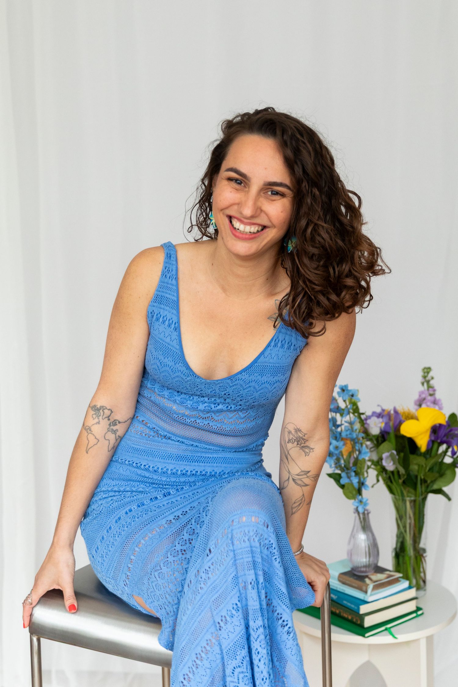 Smiling woman in blue lace dress sitting, with tattoos, curly hair, and flowers nearby.