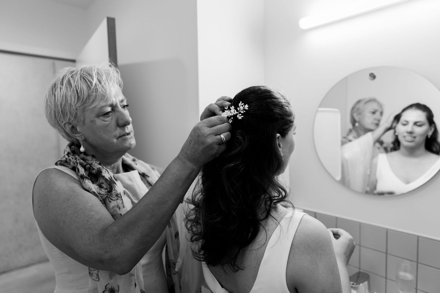 Black and white photo of older woman placing floral hair accessory on bride's dark hair, with mirror reflection in background.