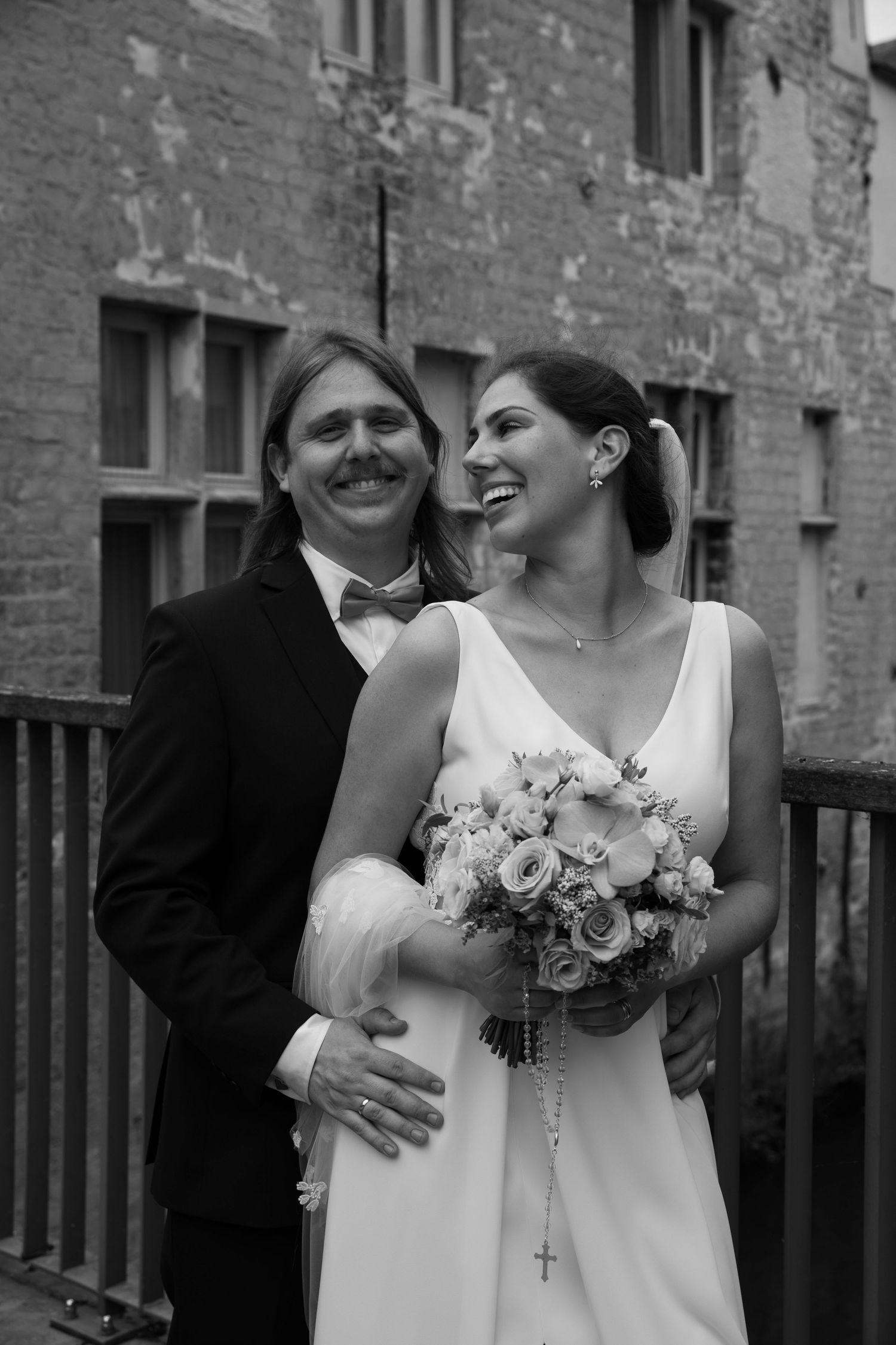 Black and white wedding photo of smiling bride holding bouquet and groom in suit with bow tie posing outdoors.