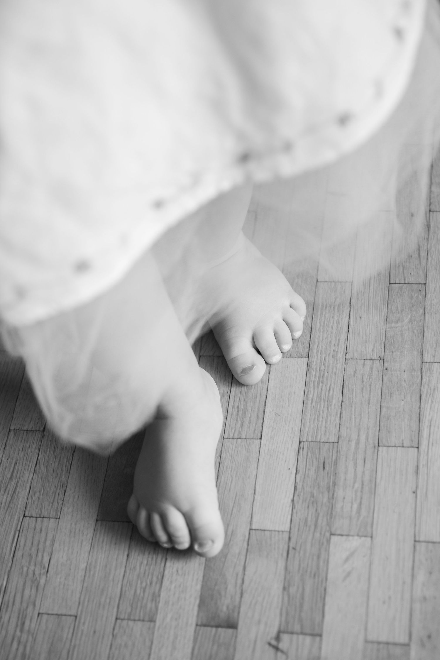 Black and white photo of tiny baby feet on hardwood floor, with white dress hem visible above.