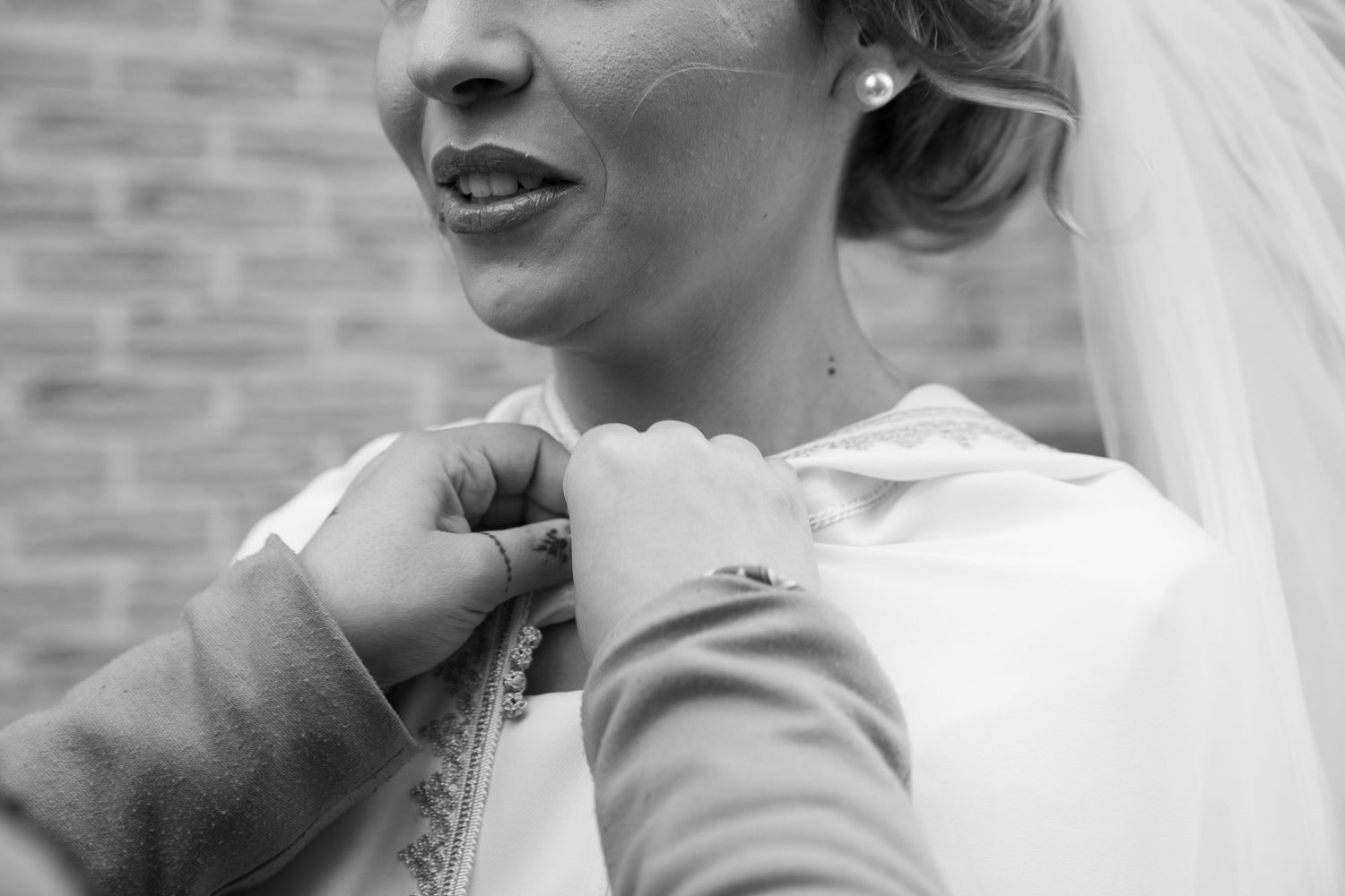 Black and white photo of a bride in white gown having her dress fastened, wearing pearl earrings and veil.