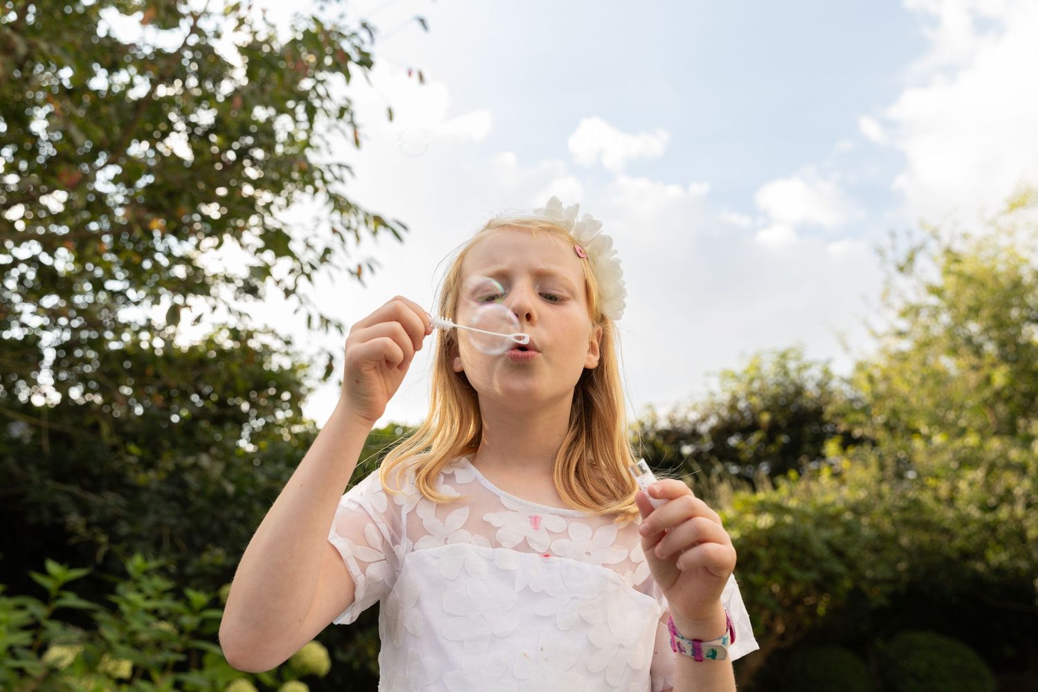 Young blonde girl blowing bubbles outdoors in a garden, wearing a white dress on a sunny day.