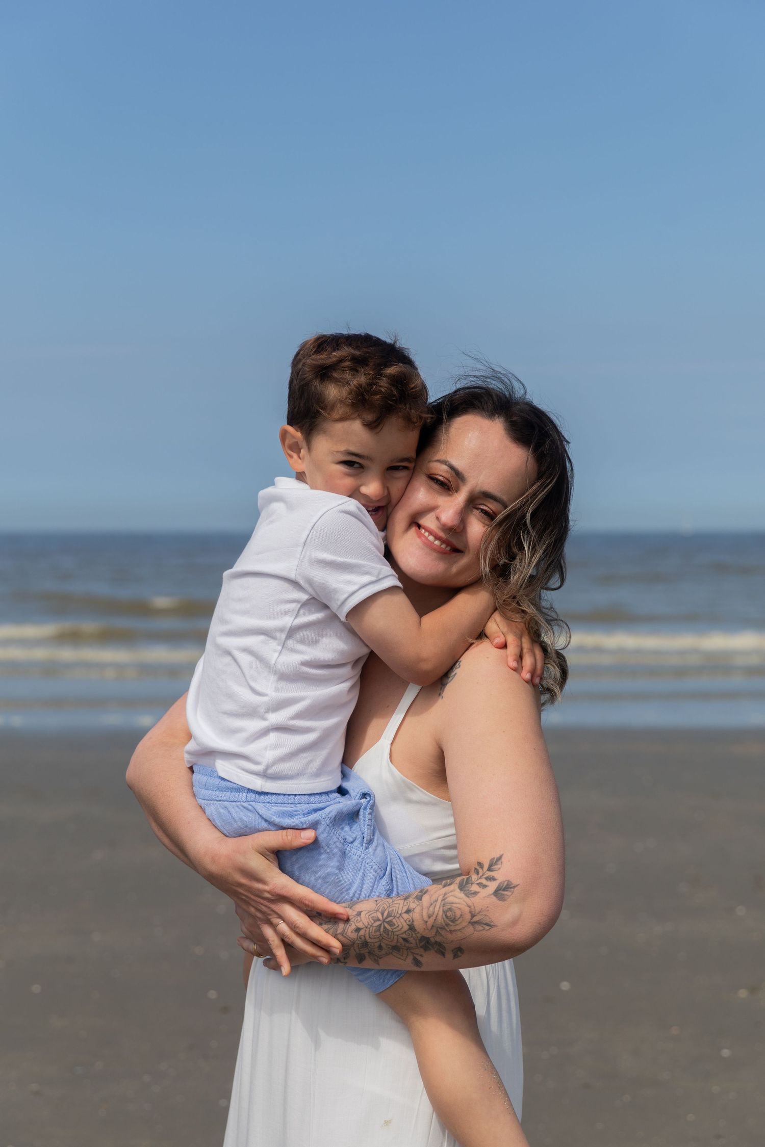 A joyful mother holds her young son in her arms on a sunny beach with ocean backdrop. photography brussels