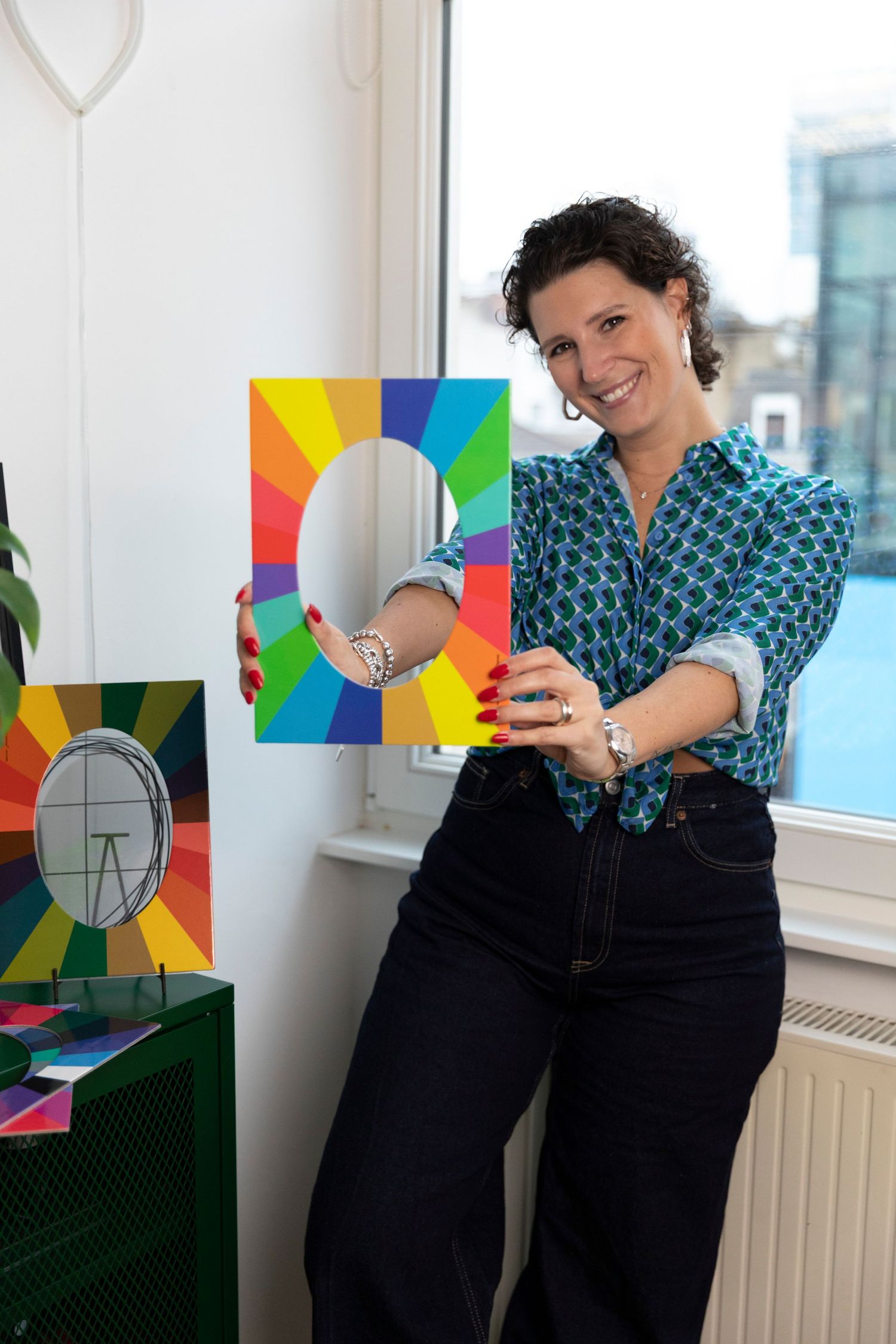 Woman in blue patterned shirt holding colorful rainbow letter O artwork, smiling in bright studio space.