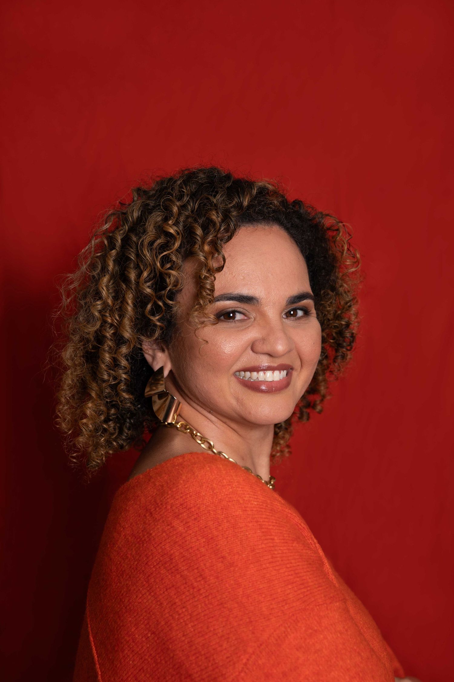 Smiling woman with curly hair wearing an orange sweater and gold necklace against a red background.