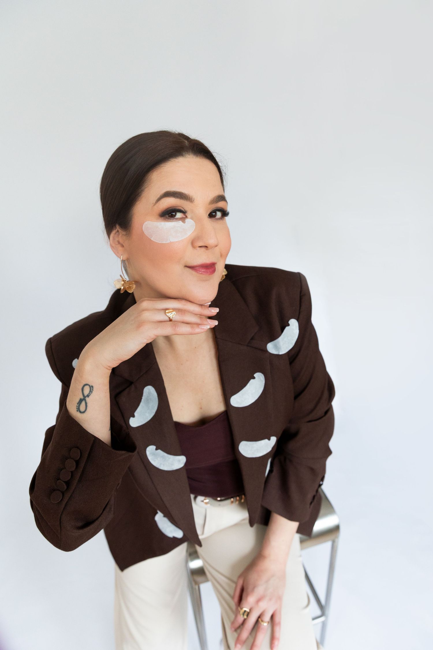 Woman in brown patterned blazer with under-eye patches poses confidently on chair against white background.
