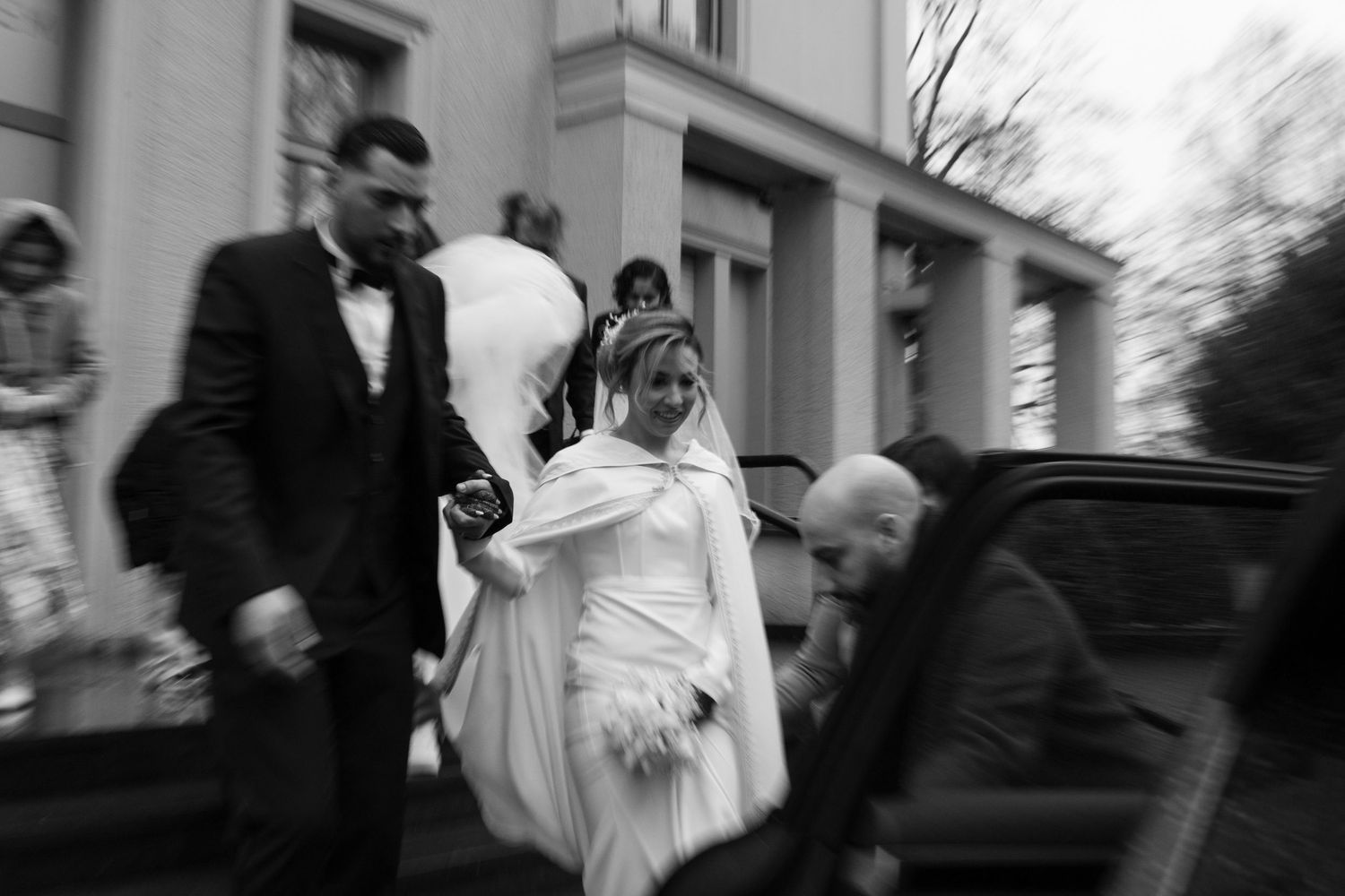 Black and white photo of bride in white fur wrap exiting building with groom and wedding guests.