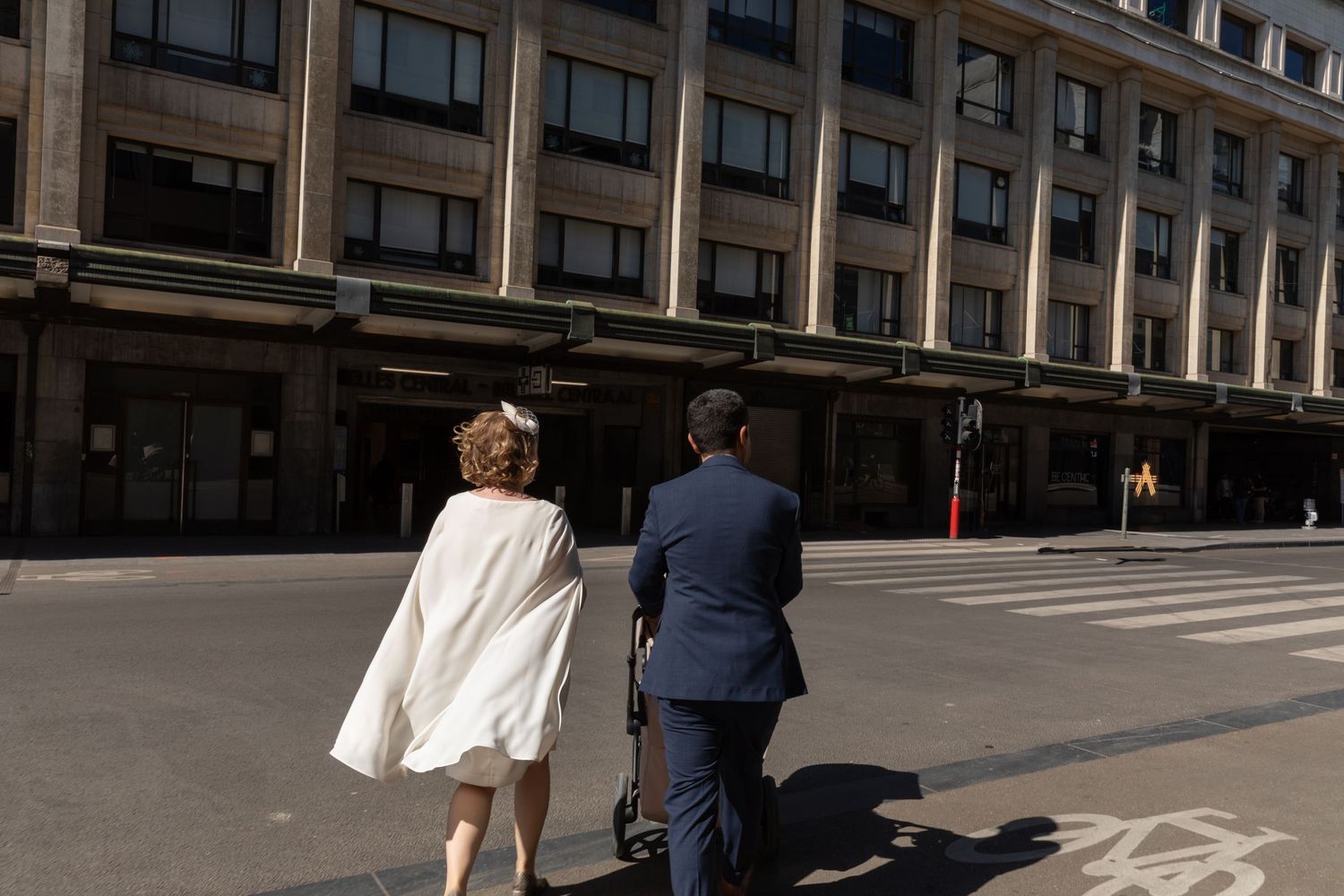 Bride and groom walking hand in hand across a sunny city street, bride wearing a white cape dress.