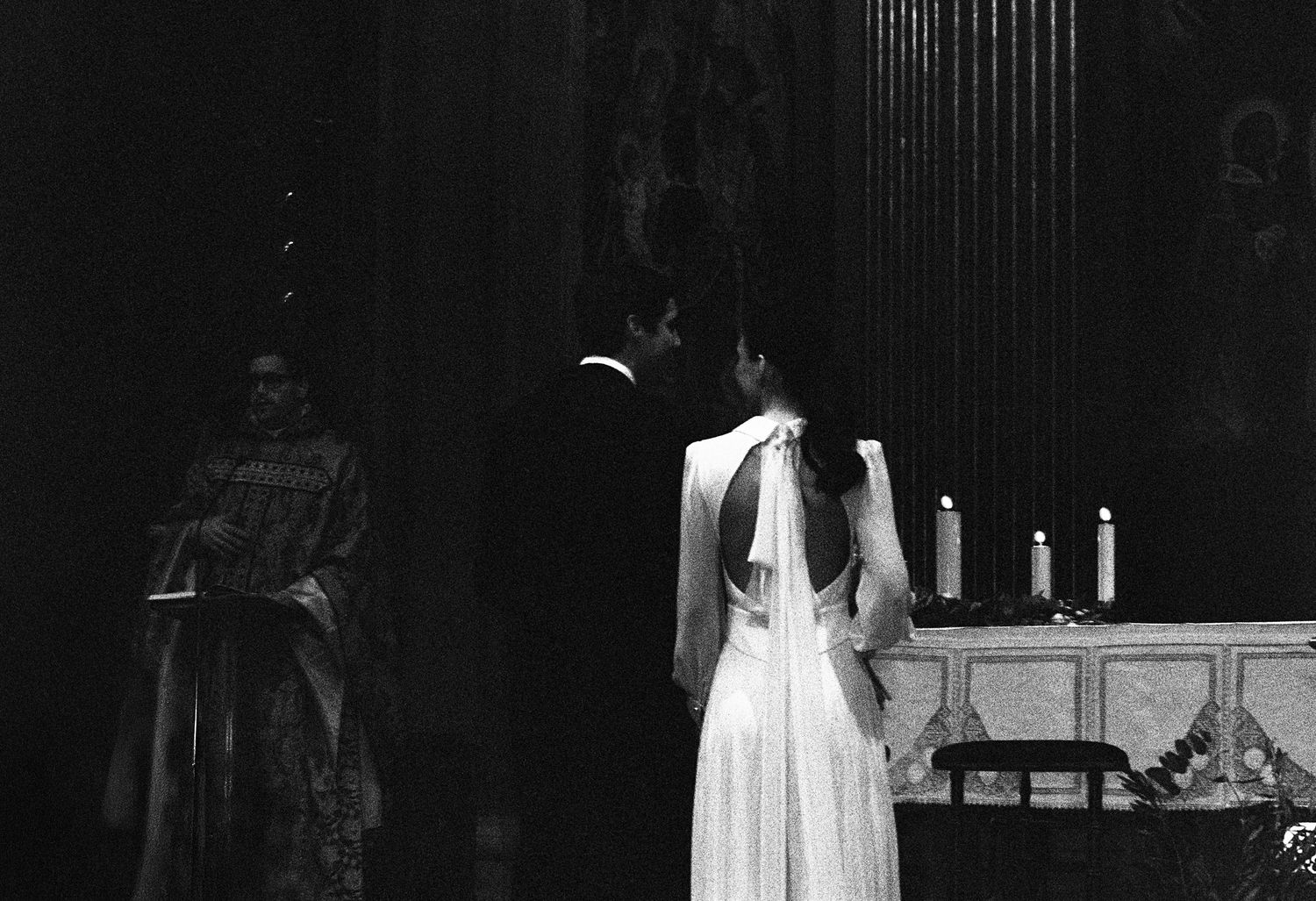 Black and white photo of a bride and groom at a church altar with candles and an officiant during a wedding ceremony.