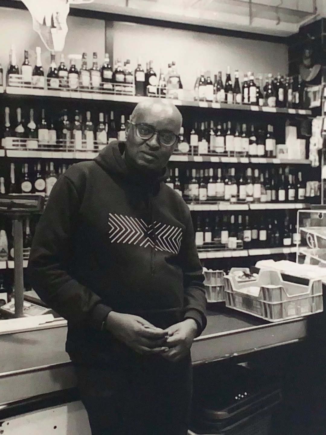 Black and white photo of a man with glasses standing behind a counter in a liquor store with shelves of bottles.