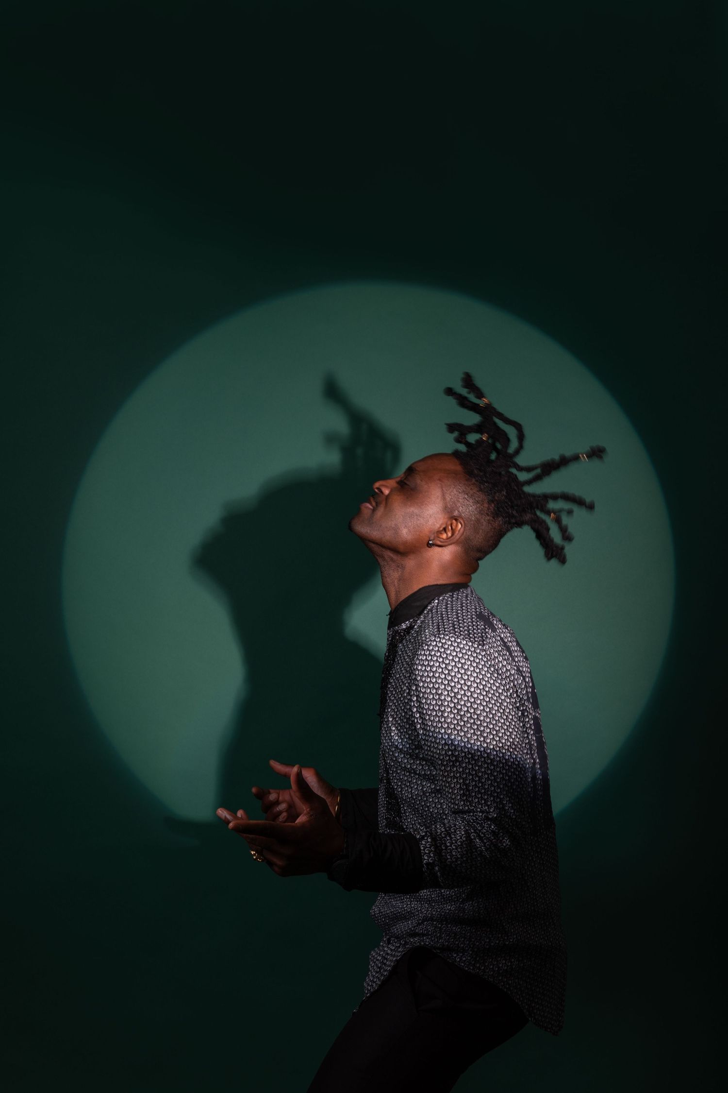 Man with dreadlocks dancing against green background with circular spotlight shadow.