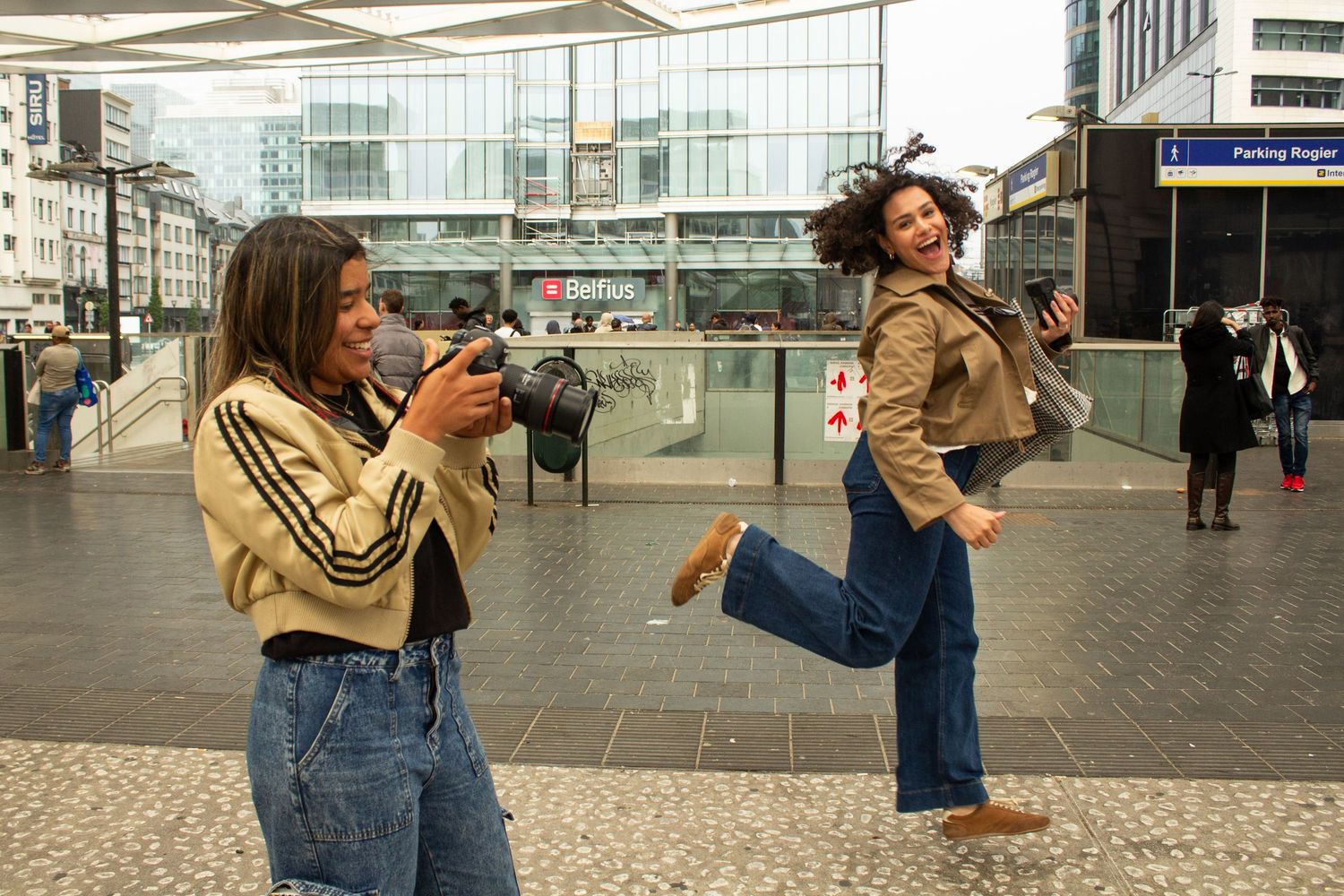 Two women in matching beige striped jackets and jeans pose outdoors, one photographing the other jumping joyfully.