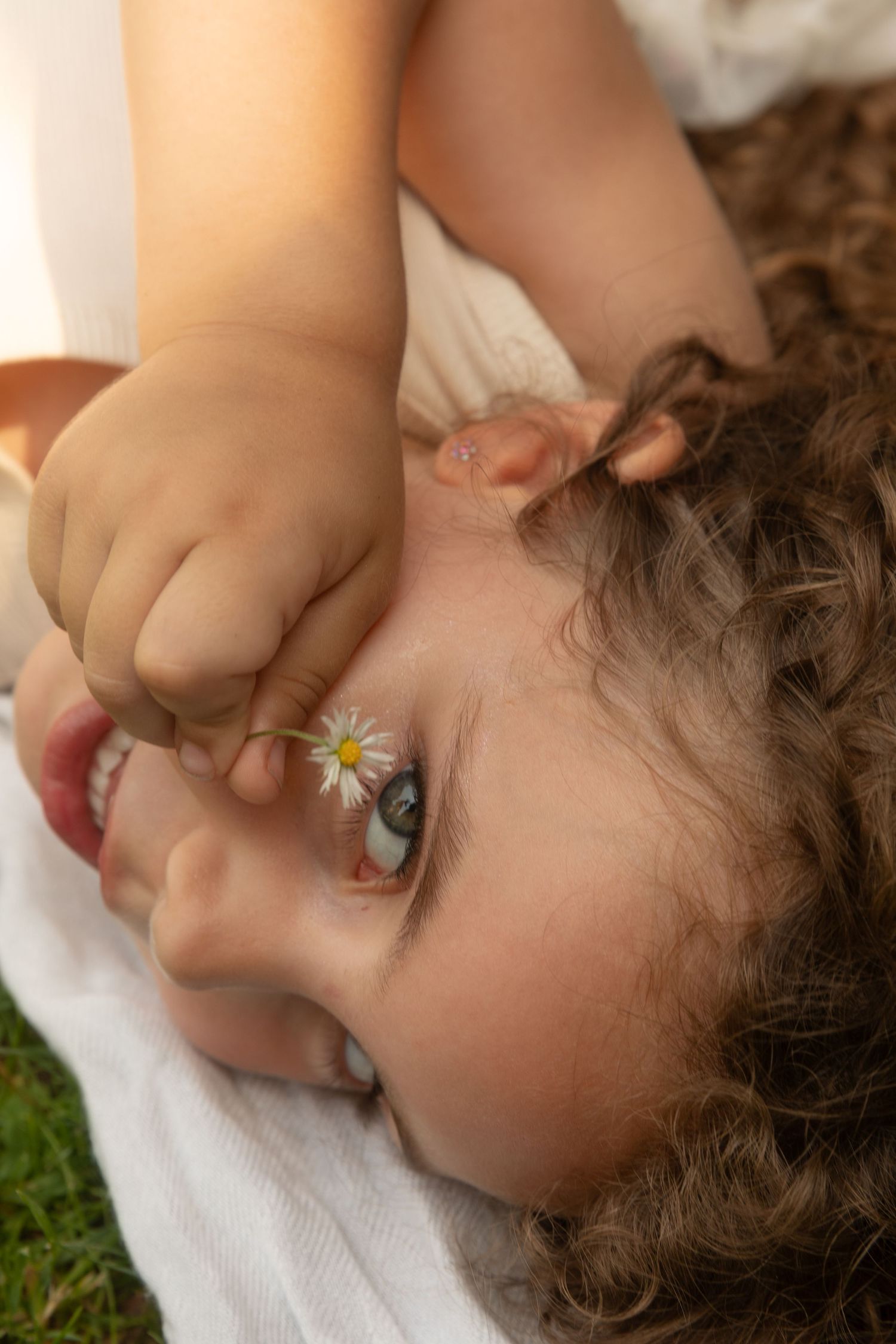Curly-haired toddler lying on grass holding a tiny daisy flower close to their eye in warm sunlight.