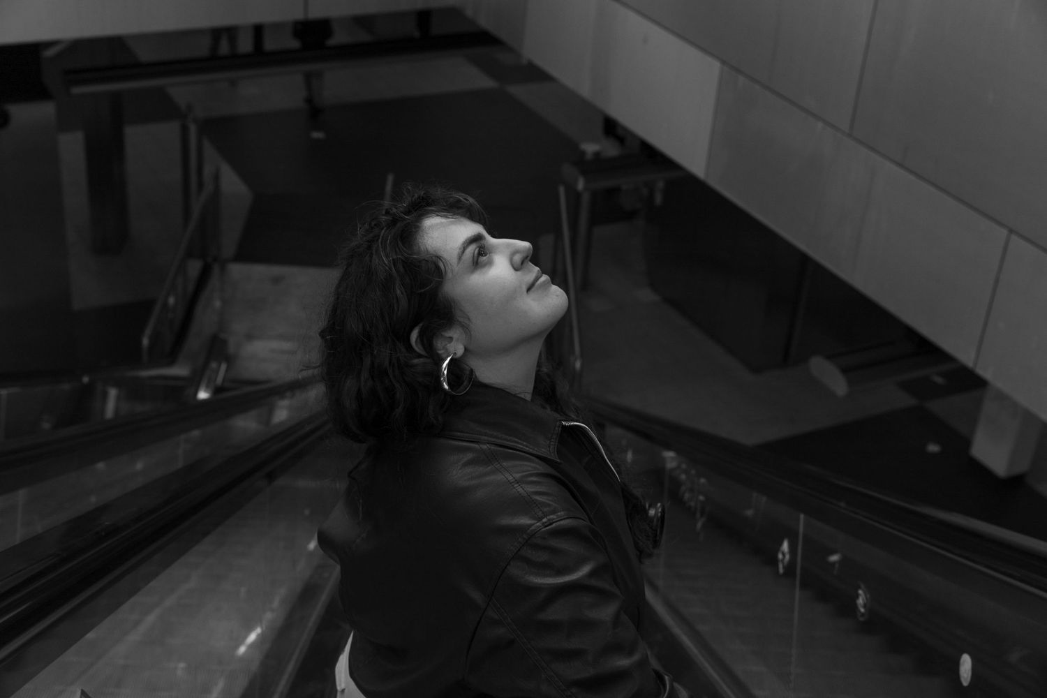 Black and white photo of a young woman in a leather jacket on an escalator, gazing upward dramatically.