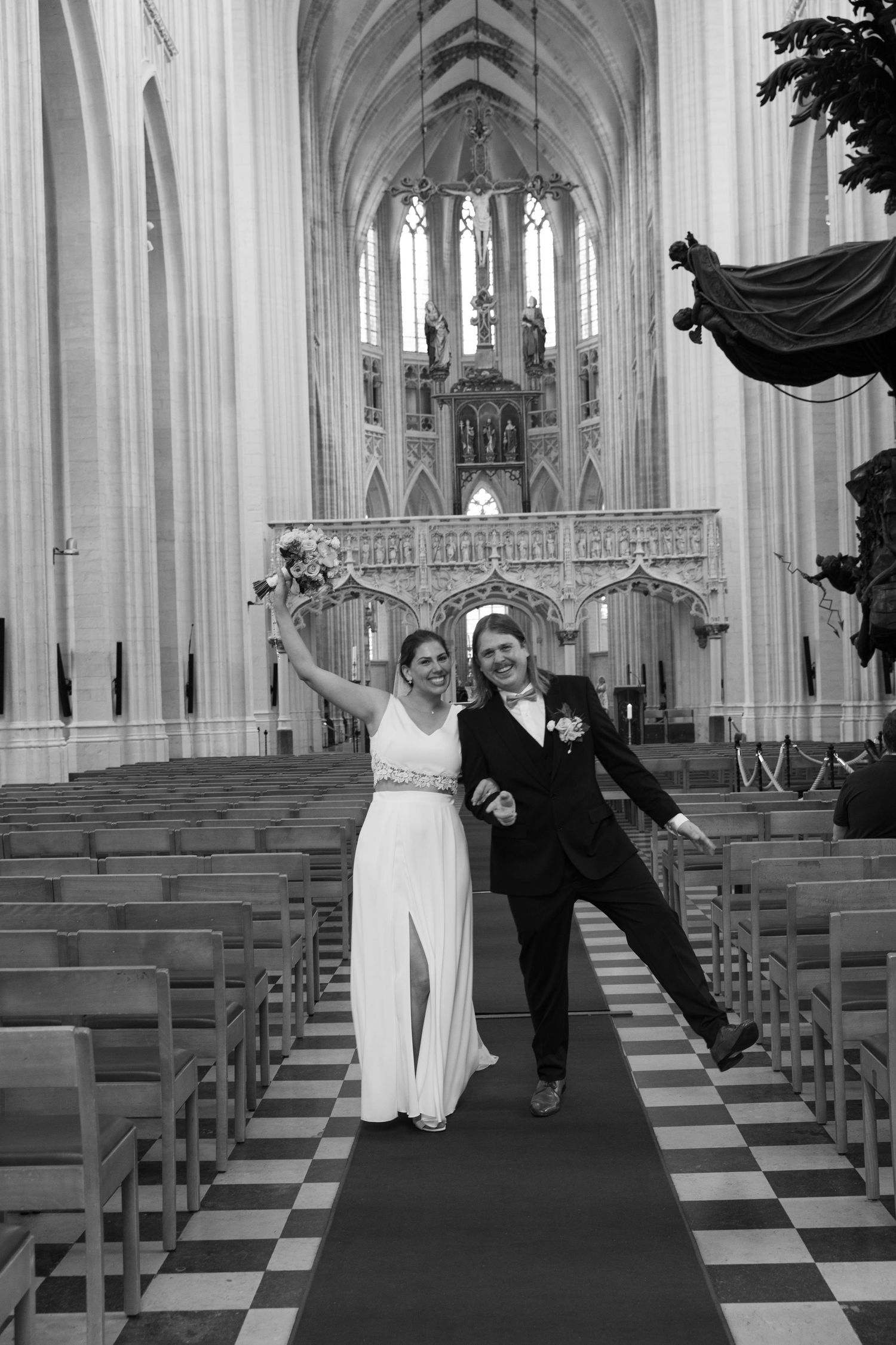 Joyful newlyweds celebrating in grand Gothic cathedral aisle after wedding ceremony in black and white.
