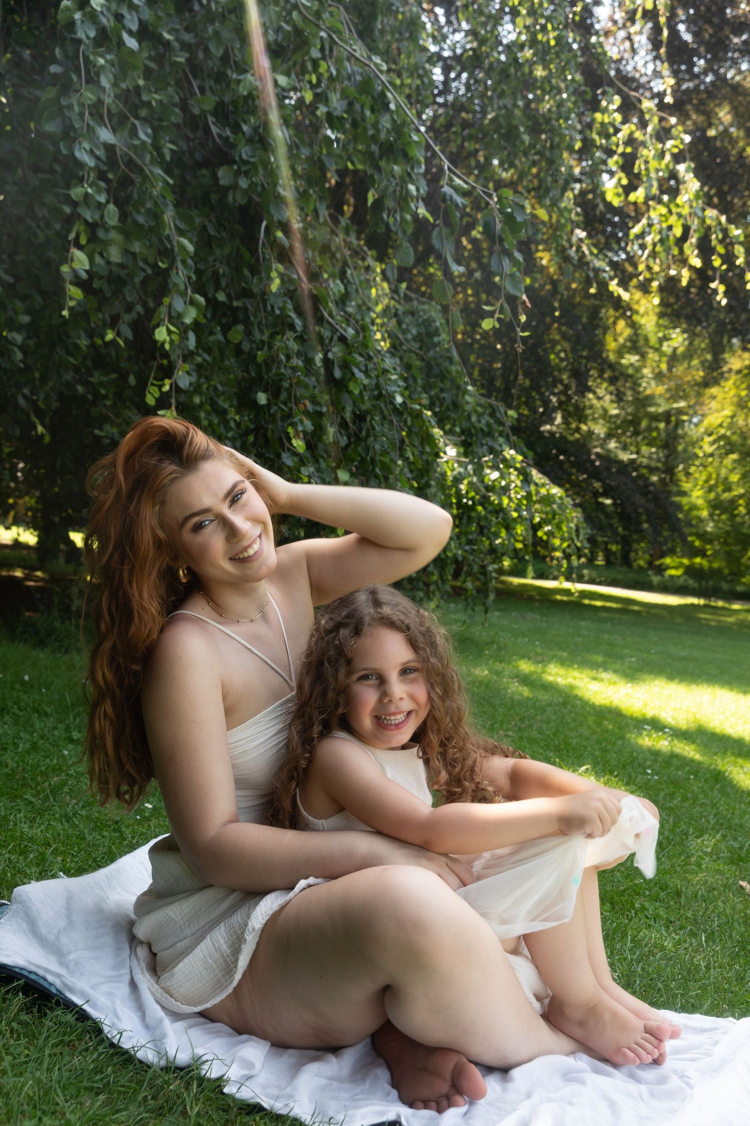 A woman and young girl in white dresses sitting on a blanket in a lush green park, smiling together.