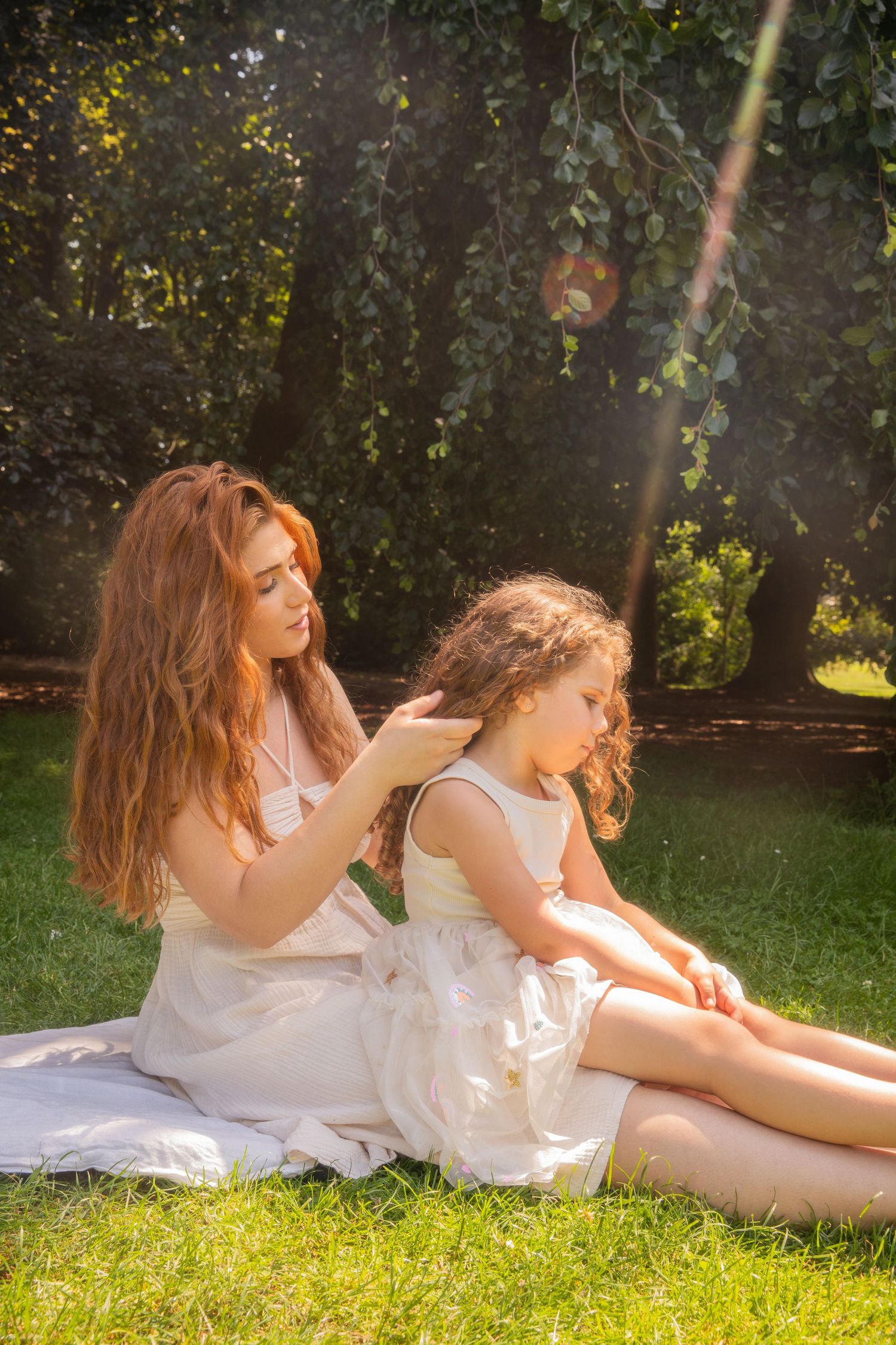 A red-haired woman in a white dress braids a young girl's curly hair while sitting on grass in a sunny park.