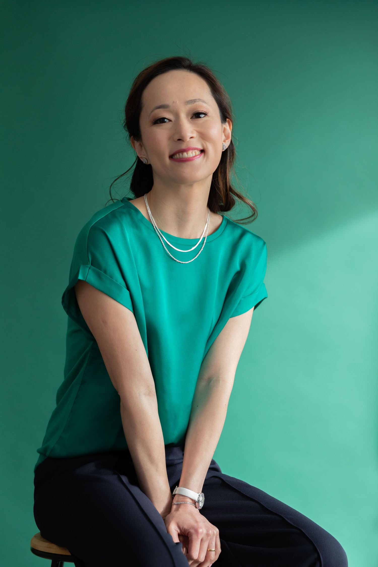 Smiling Asian woman in teal top and pearl necklace sits against green background in professional portrait.
