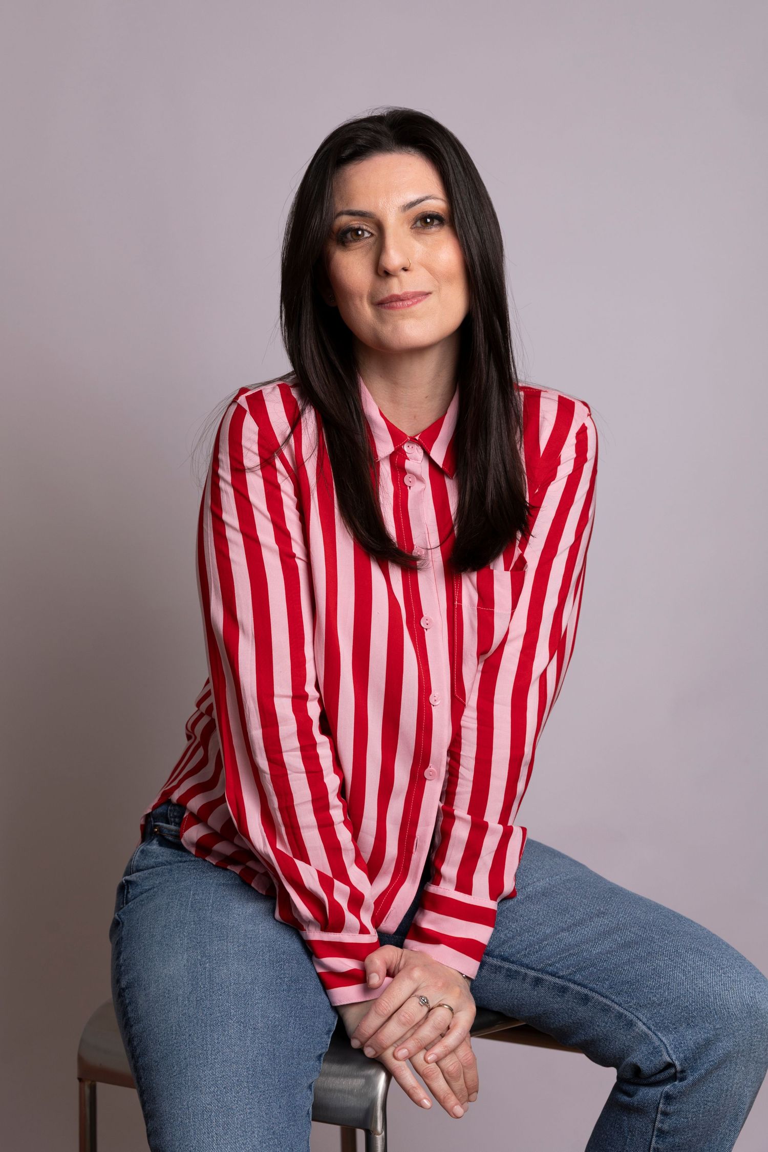 Woman with dark hair wearing red and white striped shirt and jeans, sitting on stool against gray background.