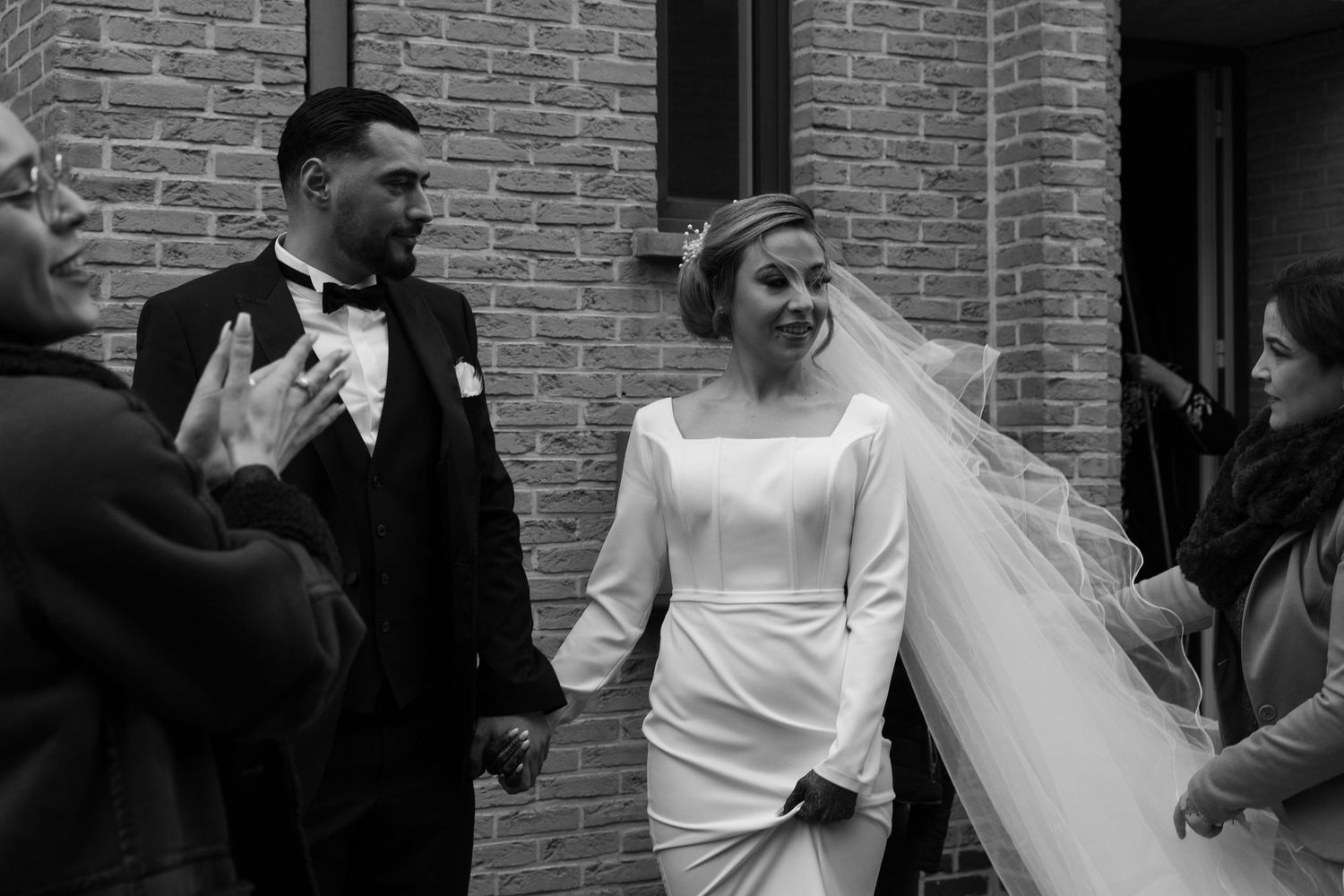 Black and white wedding photo of a smiling bride in white gown and groom in tuxedo walking hand in hand.
