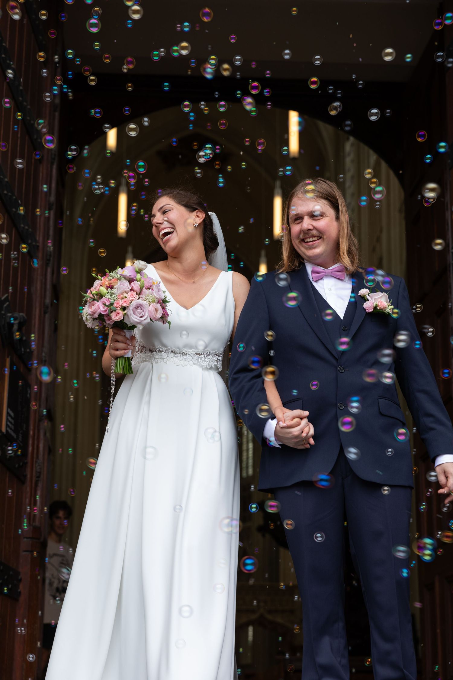 Joyful newlyweds exit church laughing as colorful soap bubbles surround them after wedding.