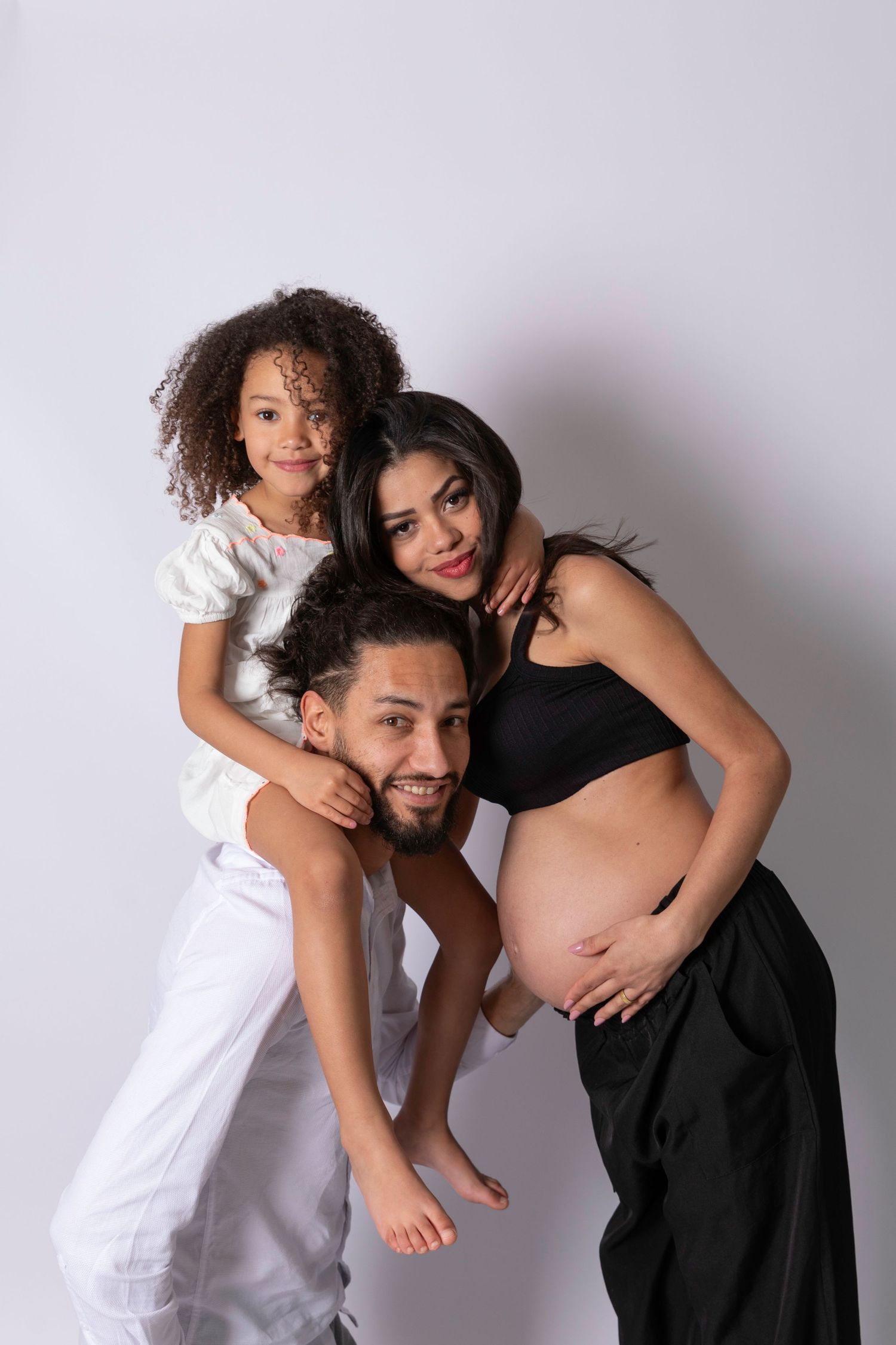 A happy family portrait with a man, pregnant woman, and young curly-haired girl on a light background.
