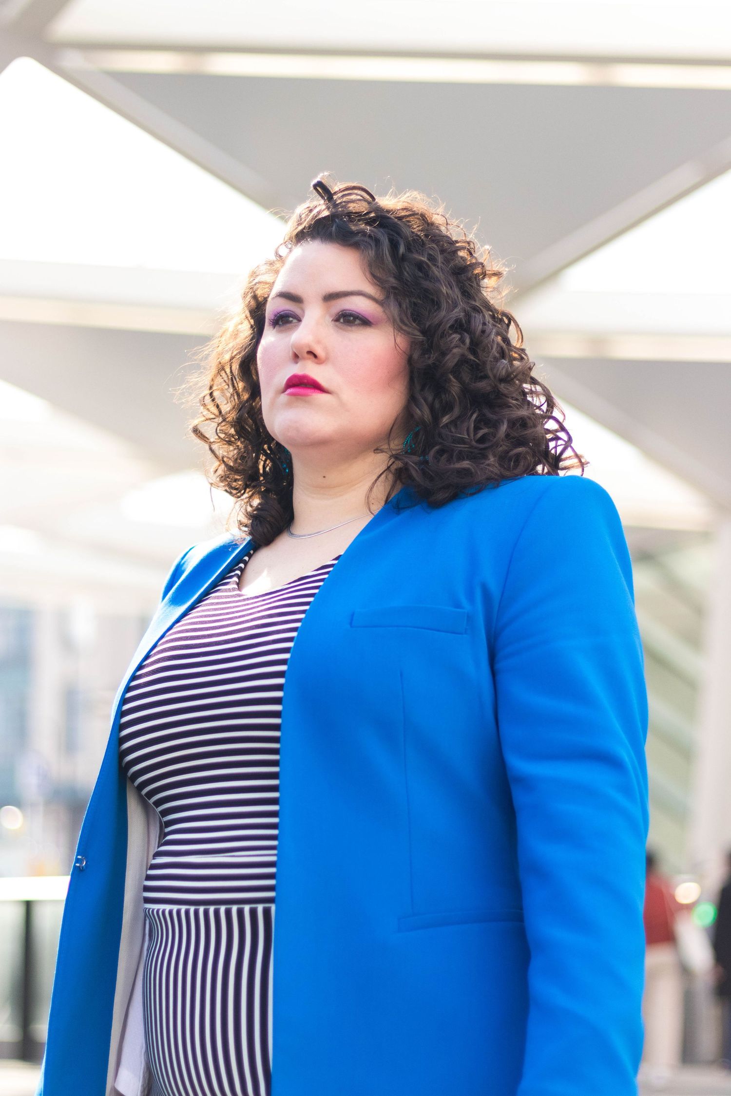 A confident plus-size woman in a blue blazer and striped dress with red lips and curly hair poses outdoors.