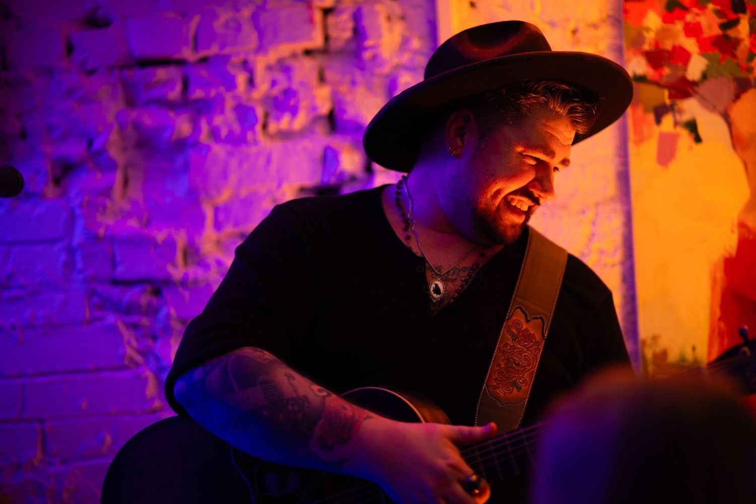 Musicians perform in dramatic purple and orange lighting on stage at an intimate venue during a live concert.