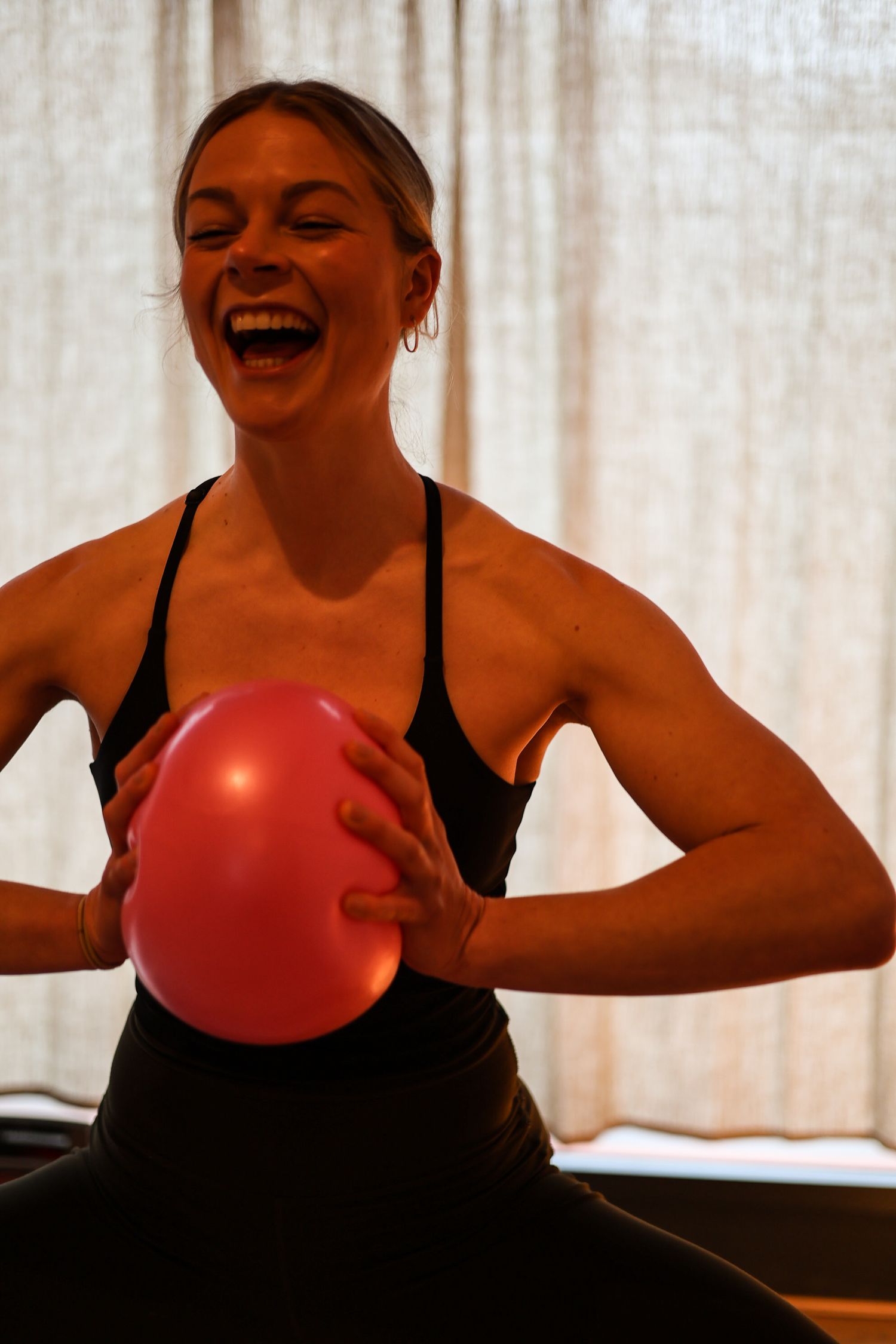 A joyful person in athletic wear holds a red exercise ball during a workout session against a light background.