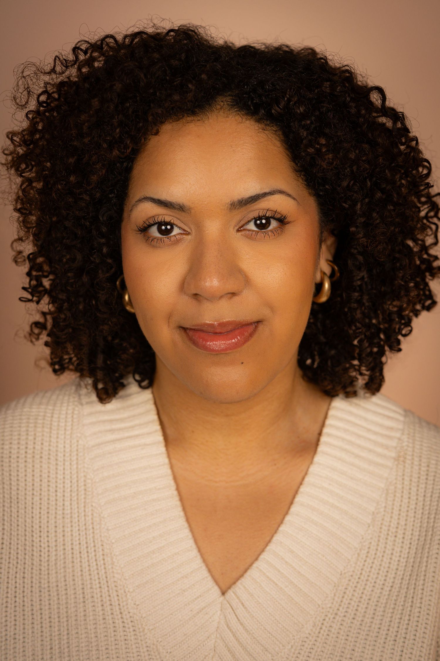 Portrait of a person with natural curly hair wearing a cream v-neck sweater against a warm brown background.