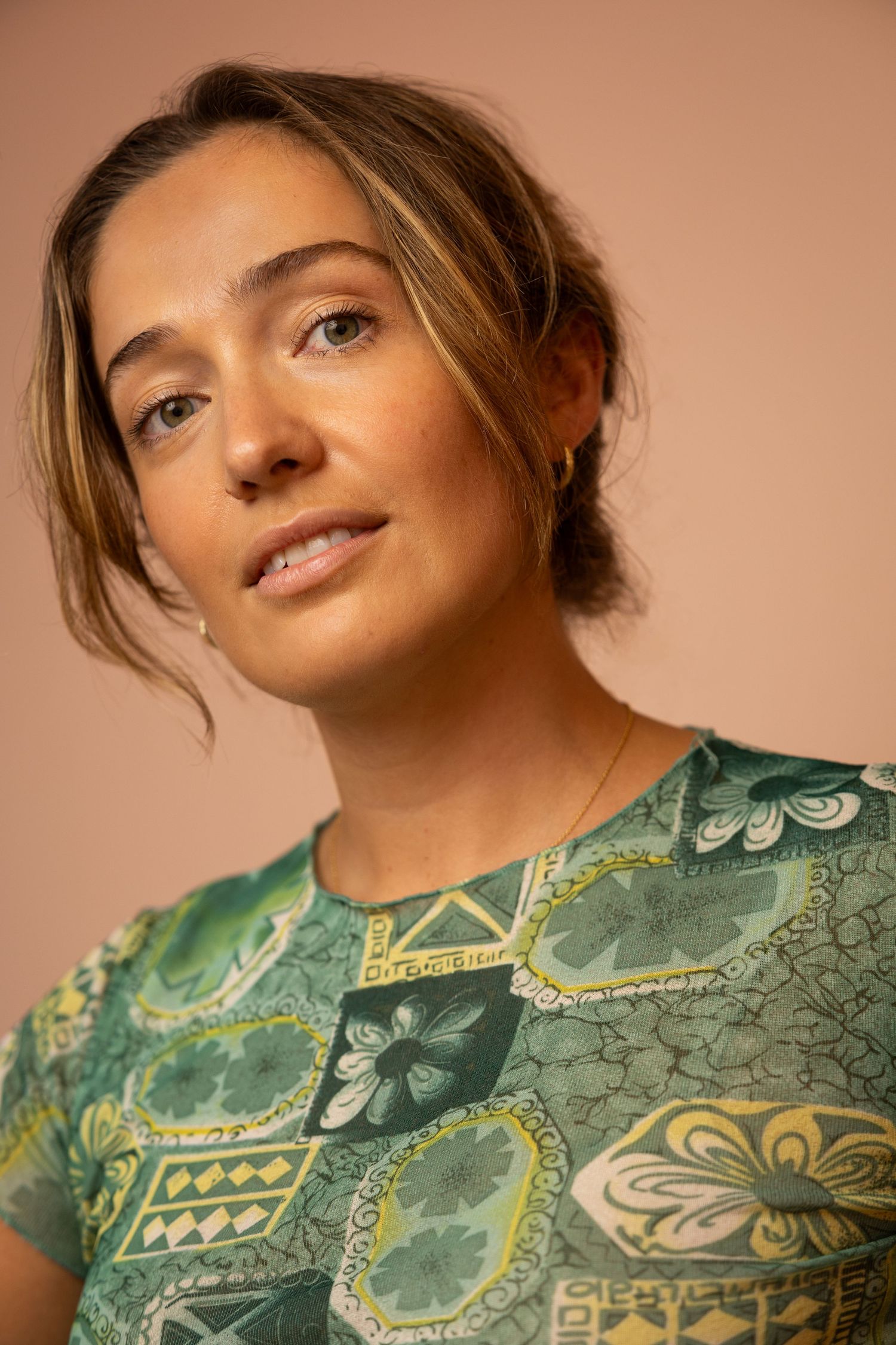 Portrait in warm lighting wearing a green floral patterned top with hair styled in a casual updo against peach backdrop.