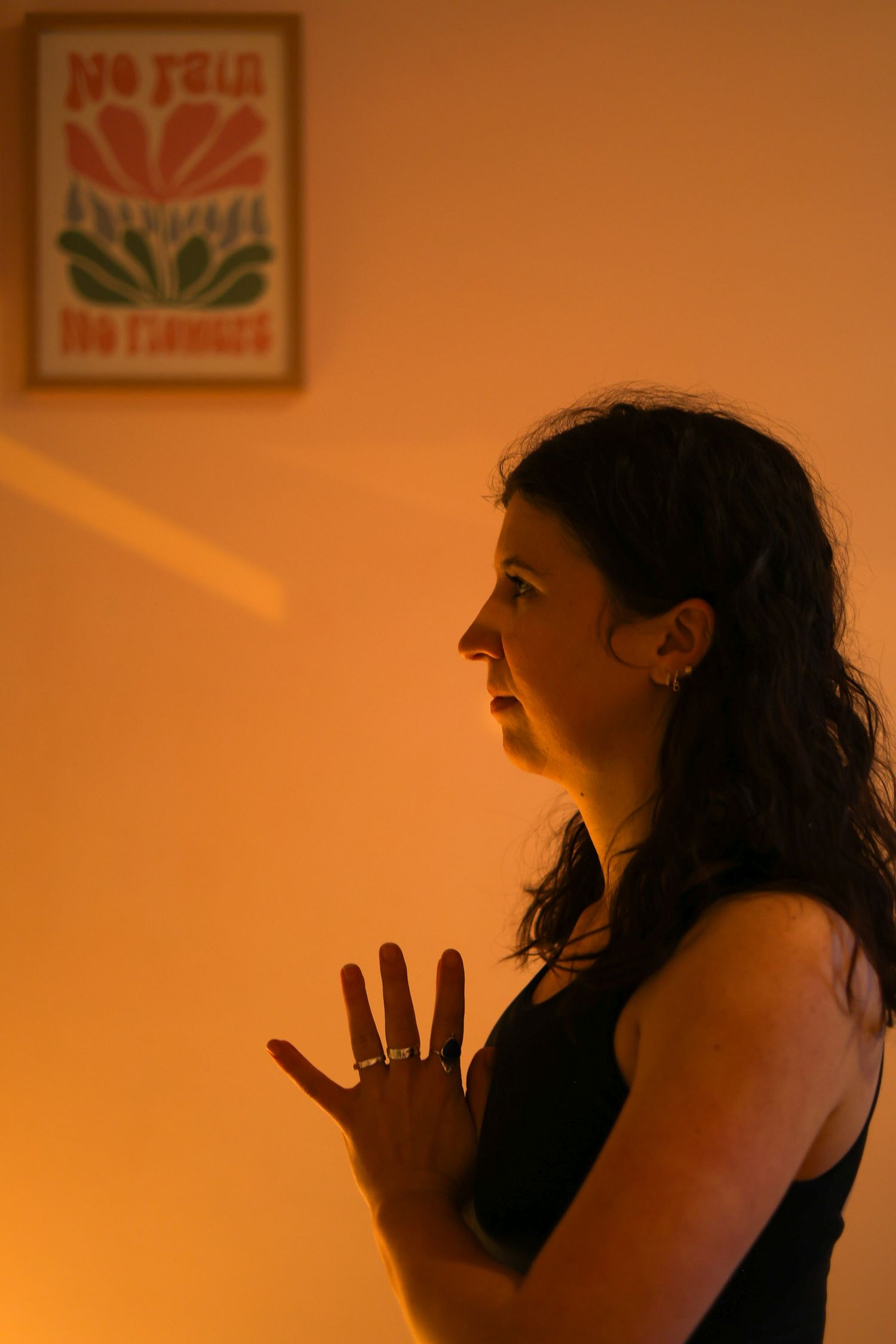 Person in dark clothing meditates with hands clasped in warm orange-lit room with art on wall.