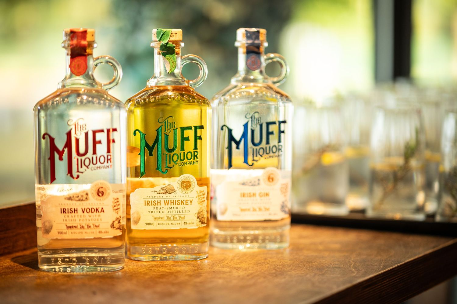 Multiple bottles of Muff Liquor Company spirits arranged in a row with warm lighting and bokeh effects in the background.