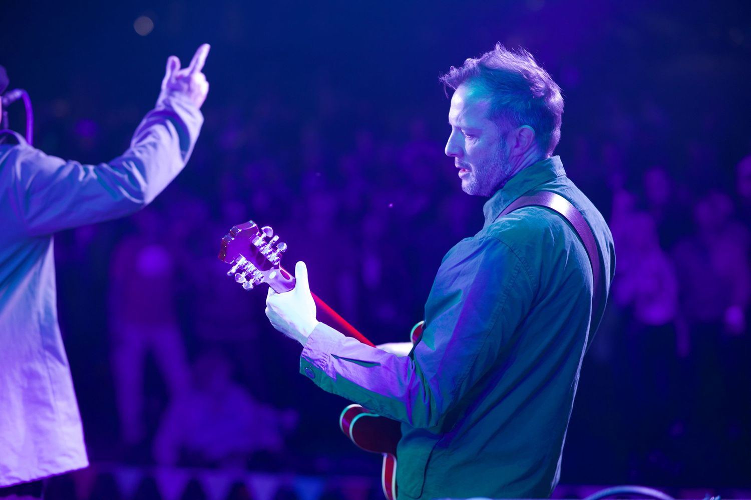 Guitarist performs on stage under purple lighting while another performer gestures to the crowd.
