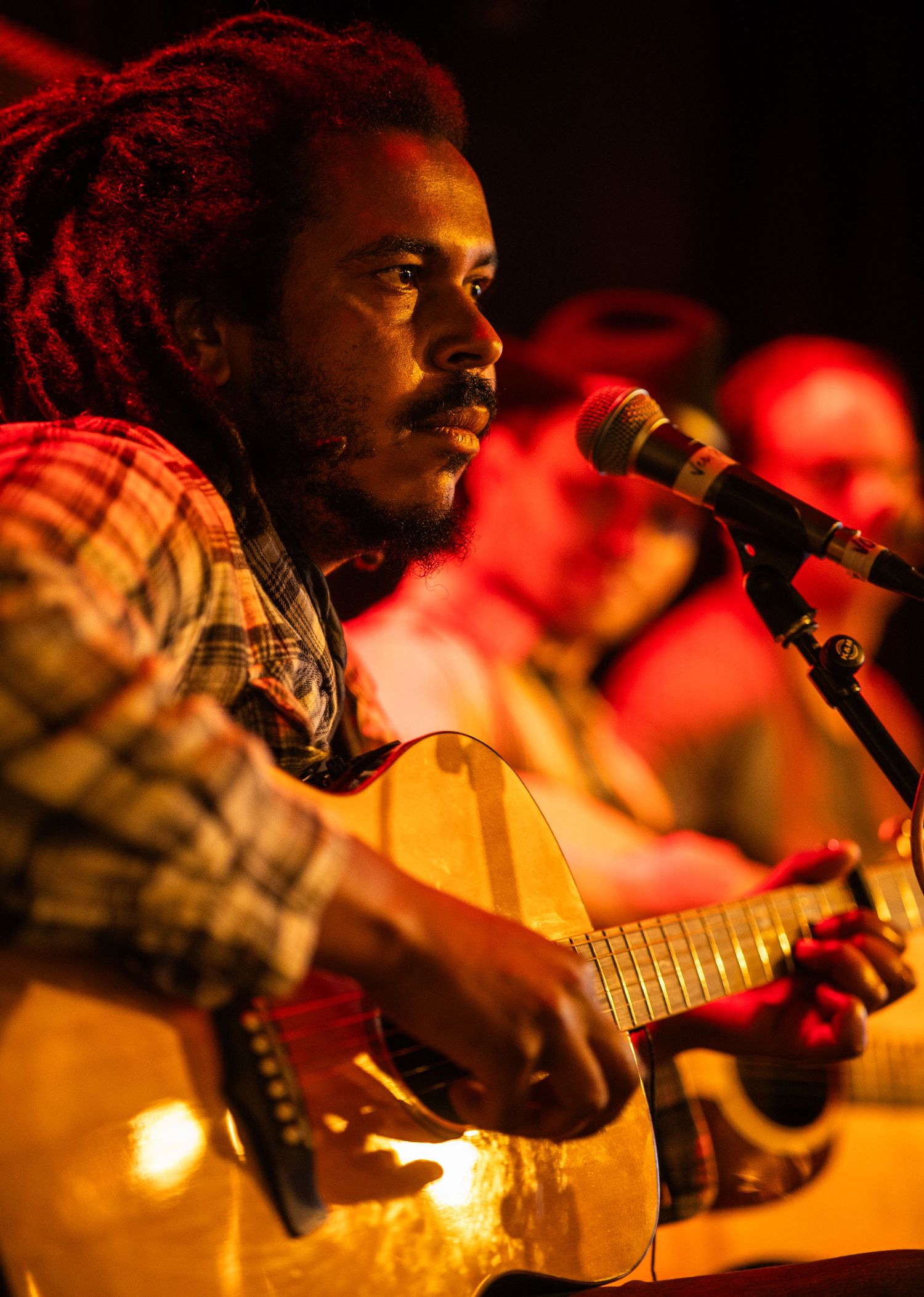 A musician performs on acoustic guitar under warm stage lighting with a red and orange ambient glow.