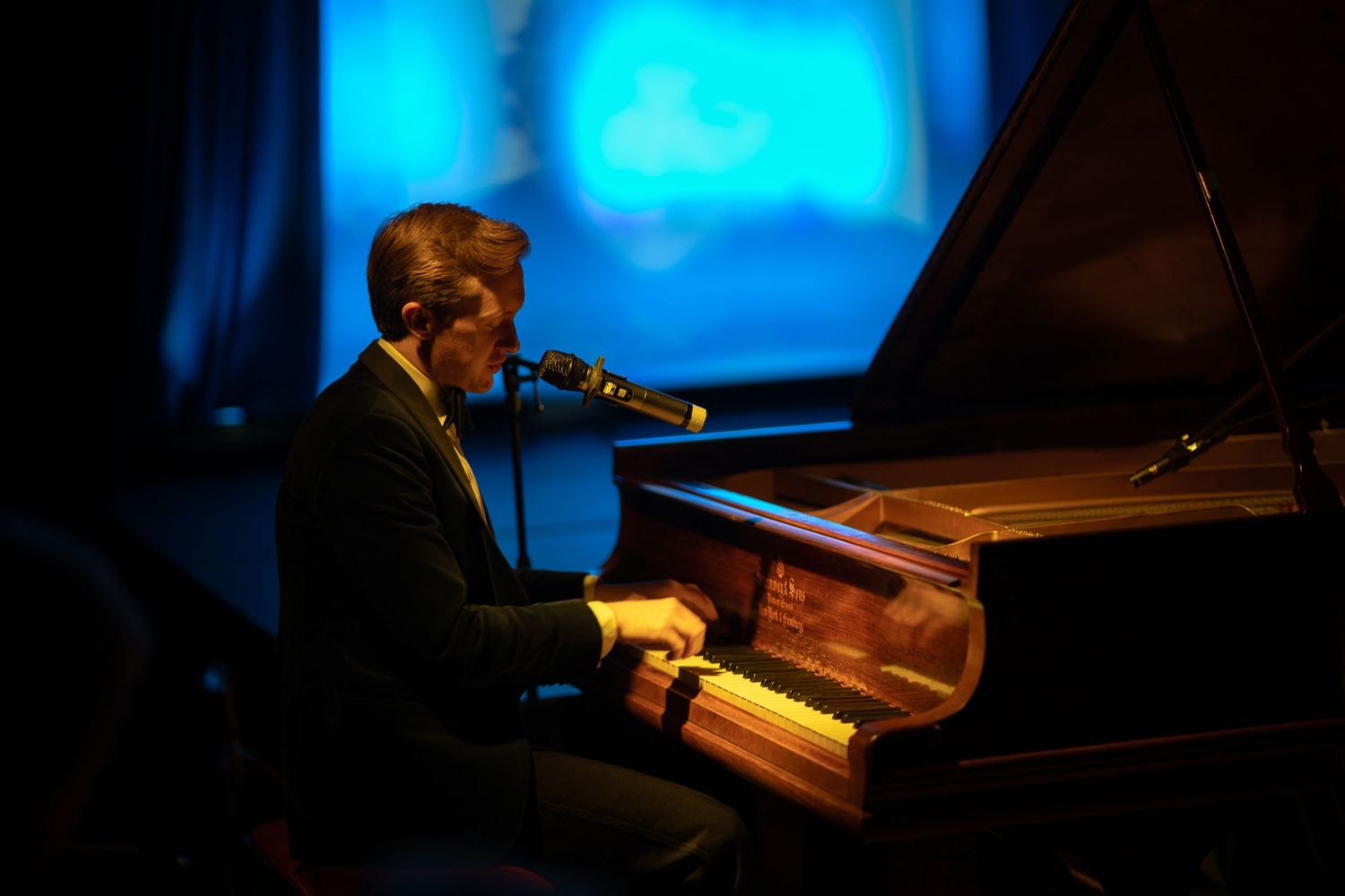 A musician performs on a piano in a dimly lit venue with dramatic blue lighting creating an atmospheric ambiance.