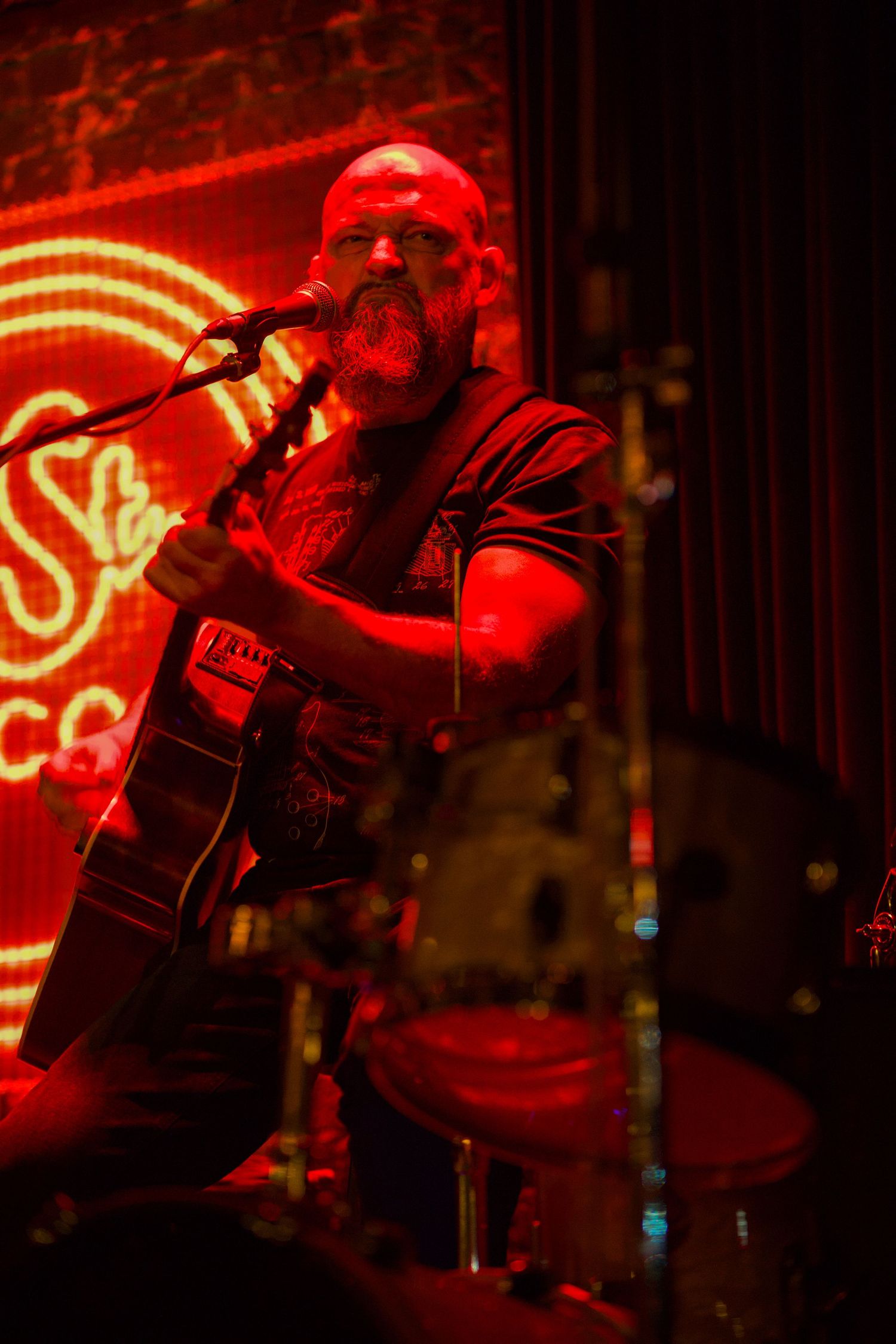 Musician performing on stage bathed in dramatic red lighting while playing guitar at a dimly lit venue.