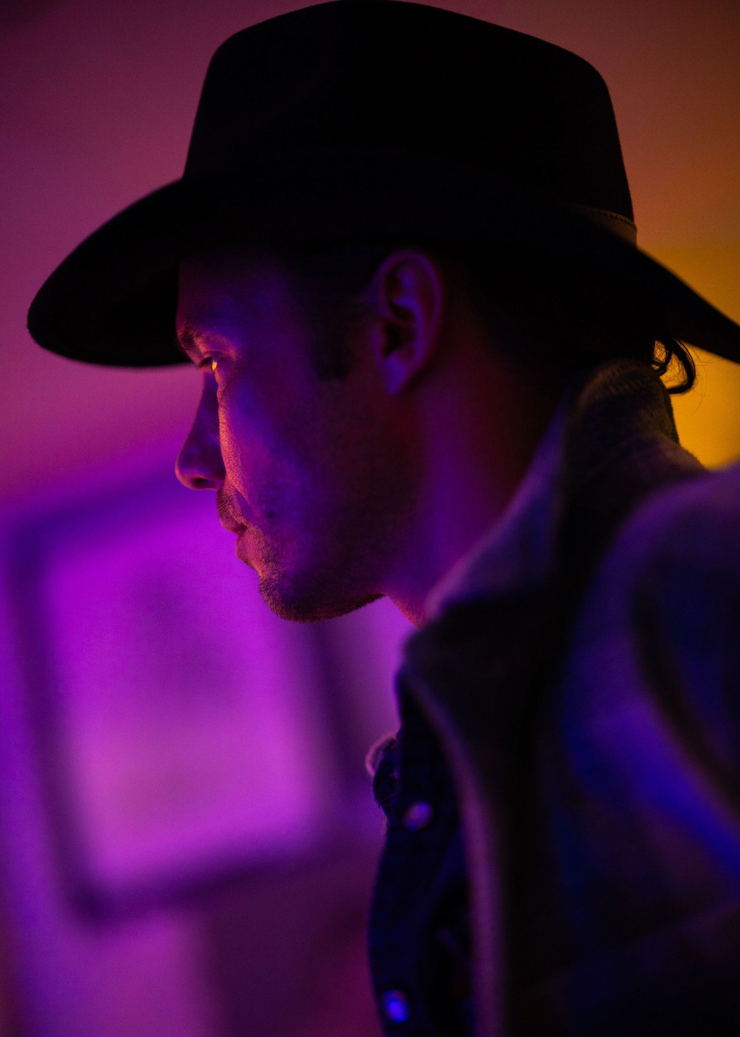 Silhouette of person in cowboy hat against dramatic purple and orange lighting in a dark moody setting.