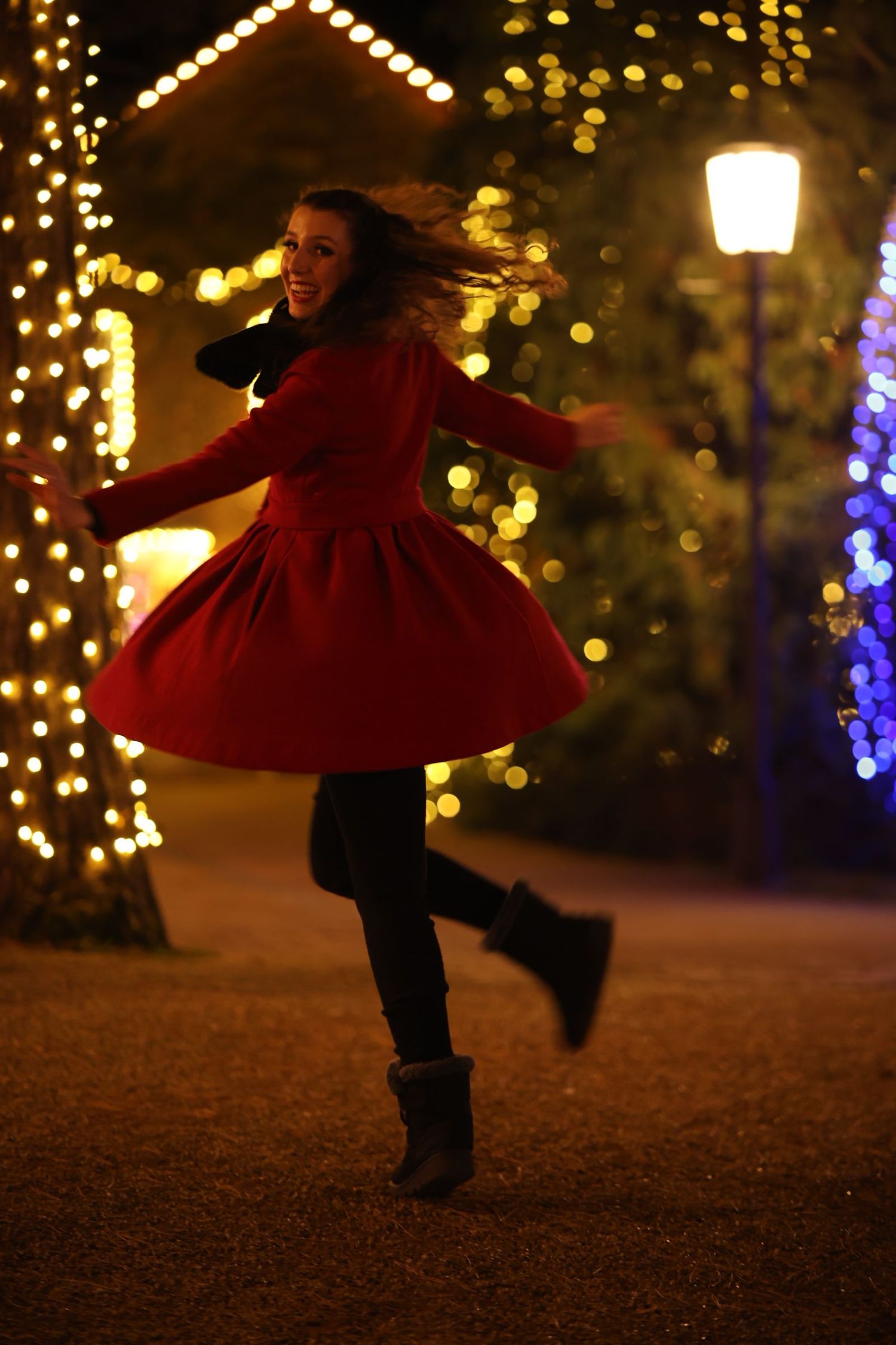 A figure in a red dress and boots twirls joyfully at night surrounded by glowing holiday lights and decorations.