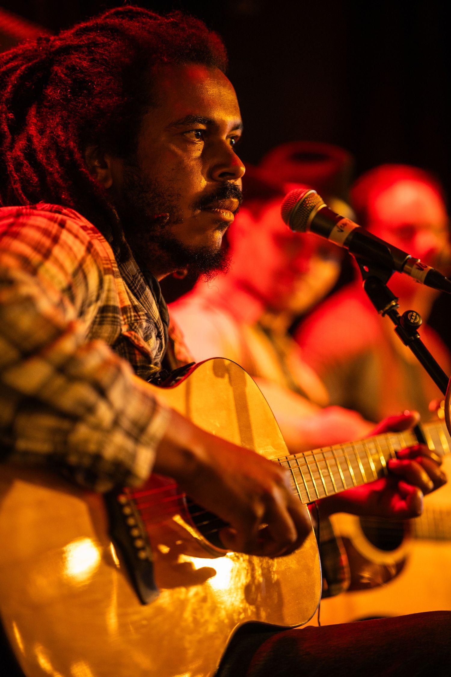 A musician performs on acoustic guitar under warm stage lighting with a reddish glow in the background.
