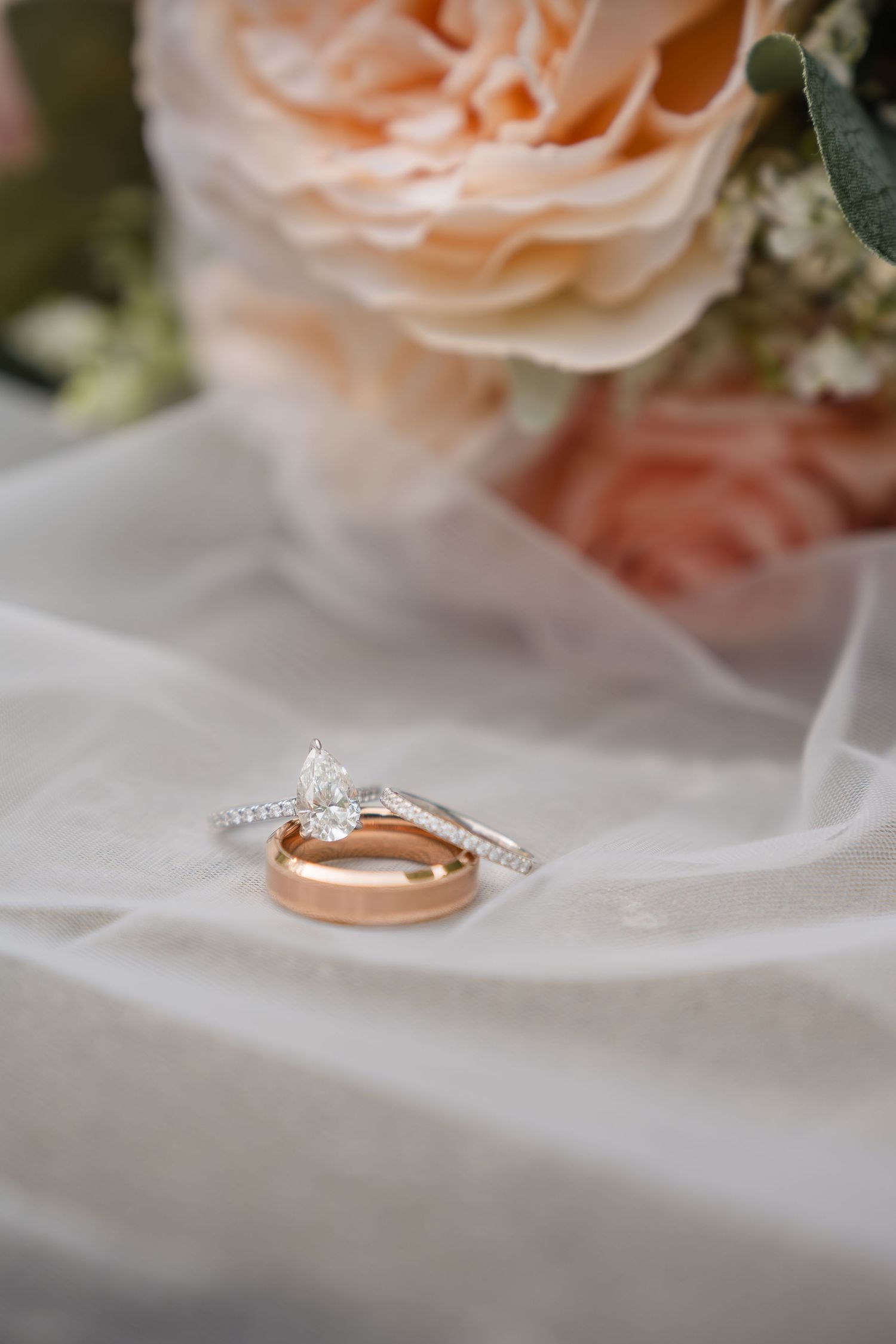 Two elegant wedding rings, one rose gold band and one diamond engagement ring, rest on white tulle with peach roses in background.