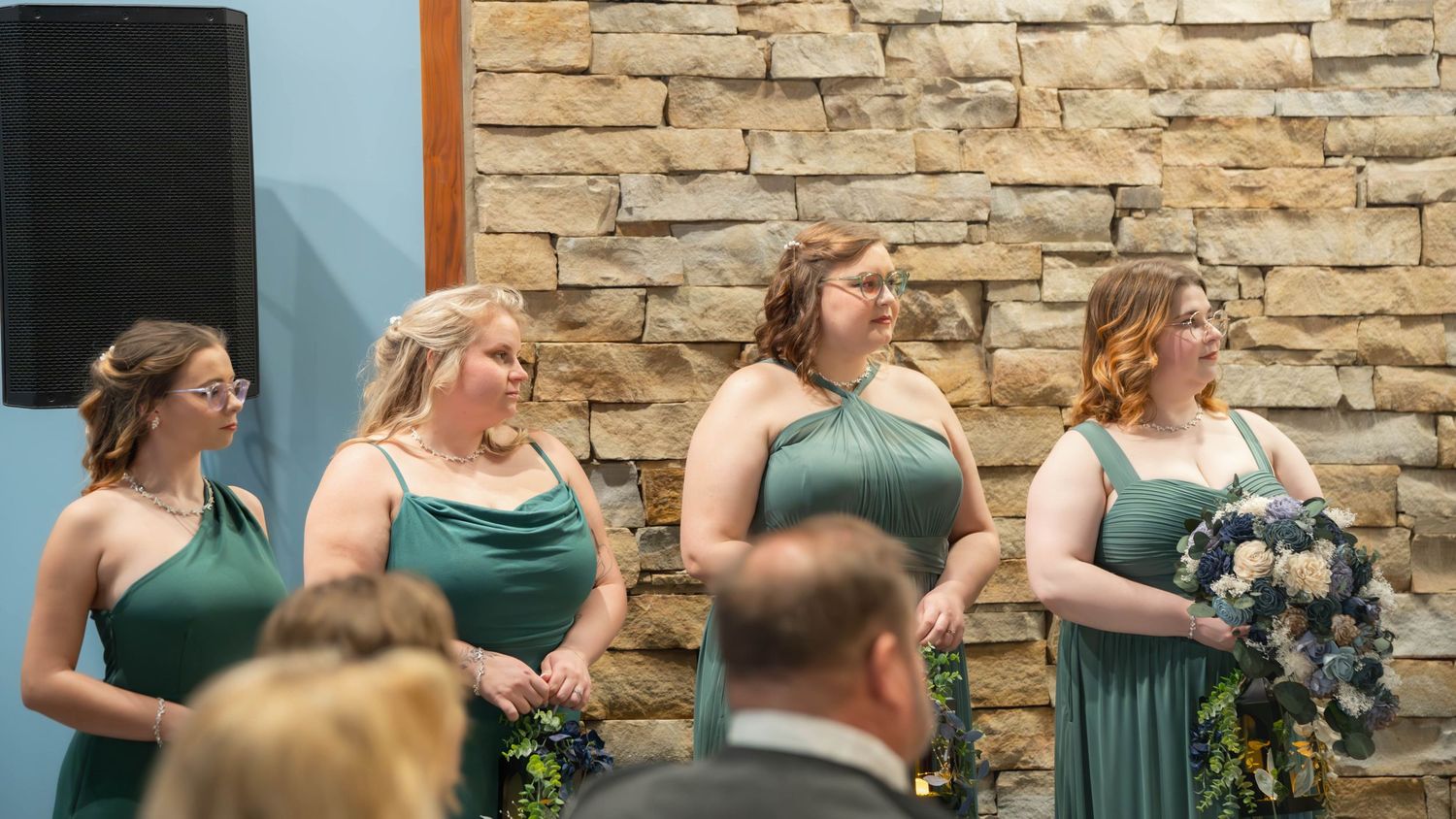 Bridesmaids wearing emerald green dresses hold bouquets while standing in front of a stone wall during wedding ceremony.