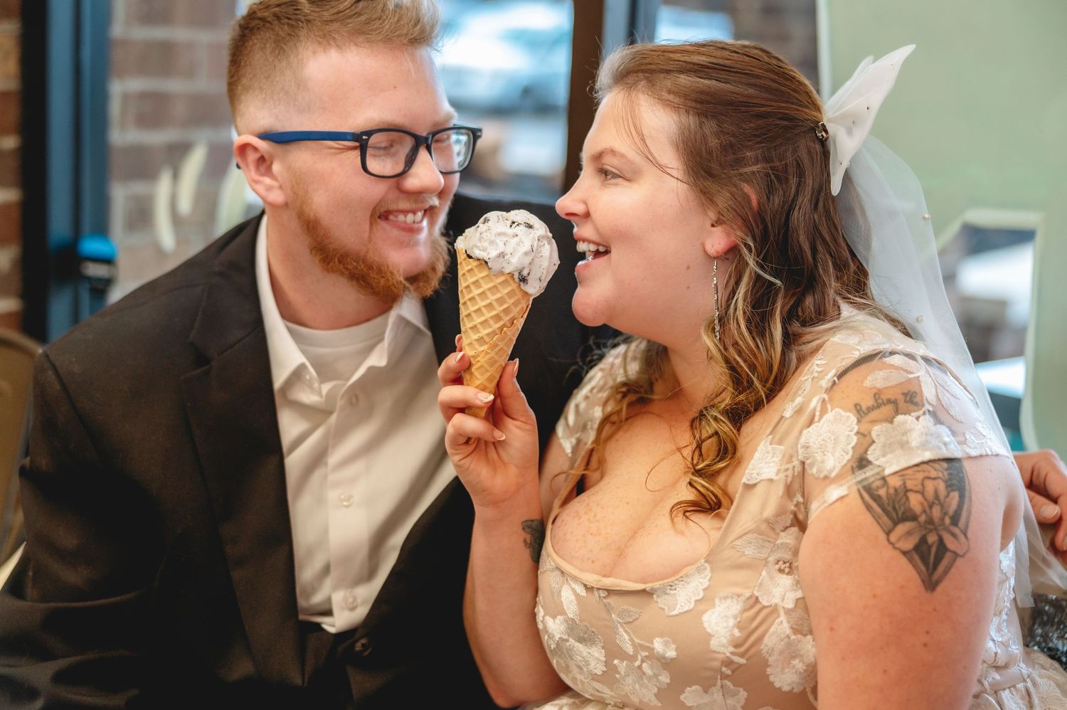 A couple dressed in wedding attire share a joyful moment while enjoying ice cream at an indoor venue.