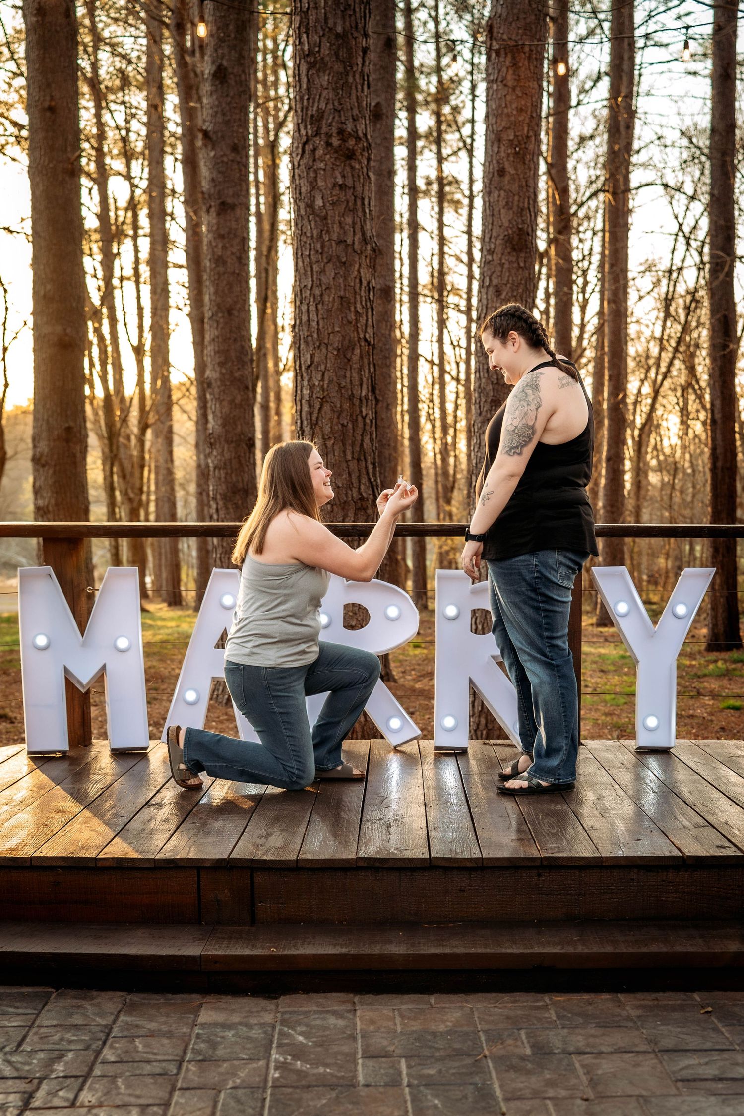 A romantic proposal moment on a wooden deck in a sunlit forest with decorative white letters spelling 'MARRY' in the background.