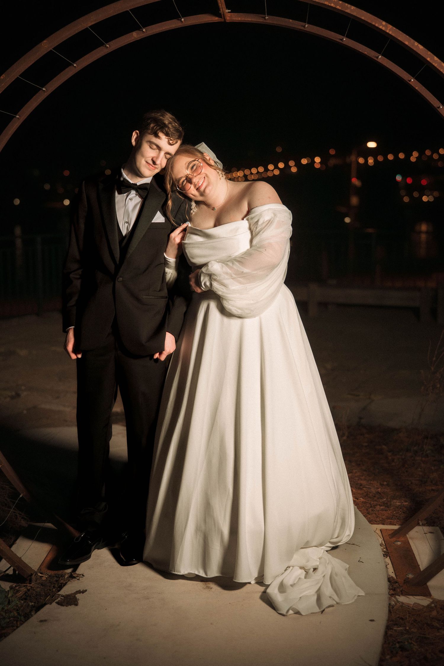 Couple poses in formal wedding attire at night under an archway with twinkling lights in the background.