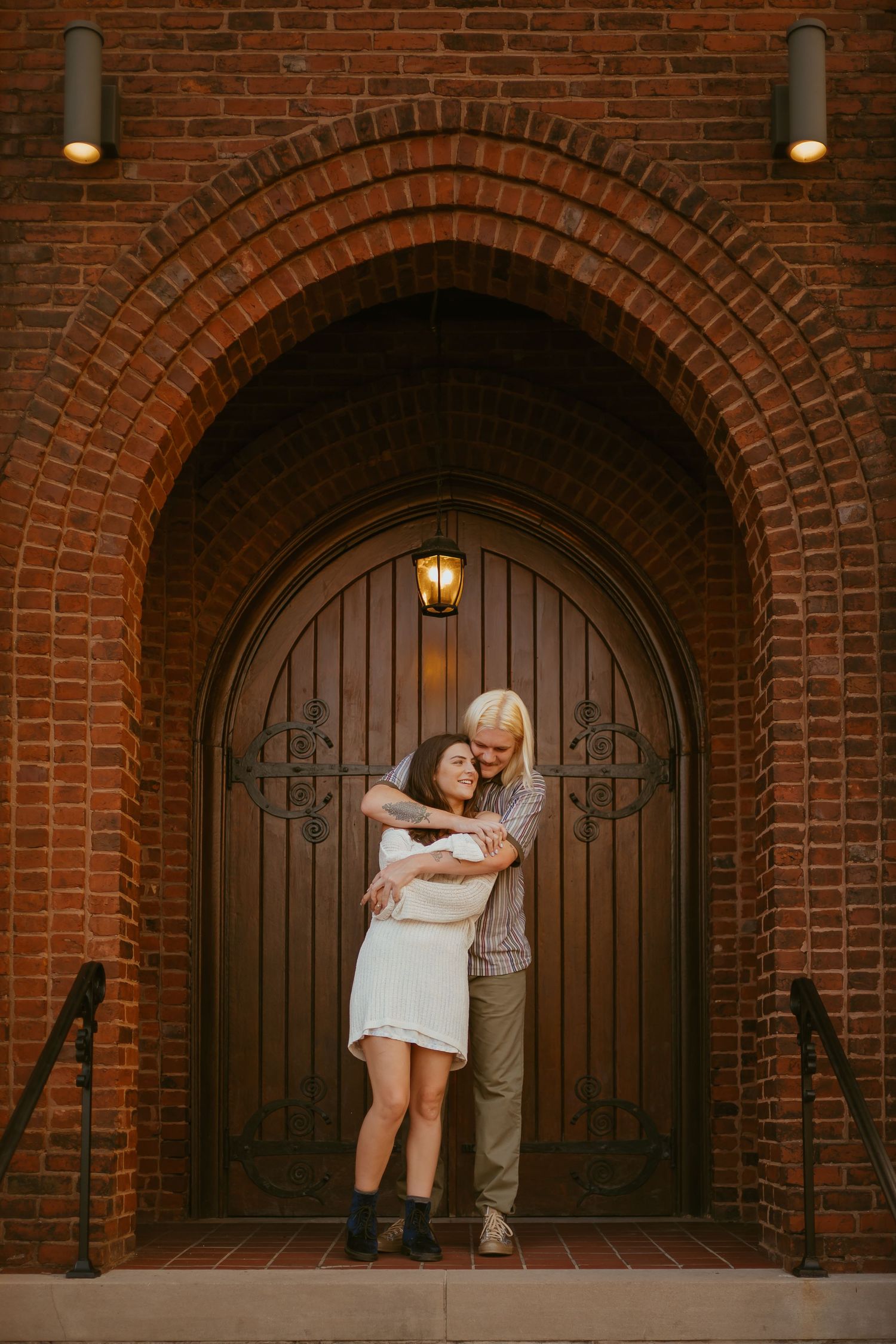 Couple poses for engagement photos in front of rustic arched brick doorways with warm lighting at sunset.