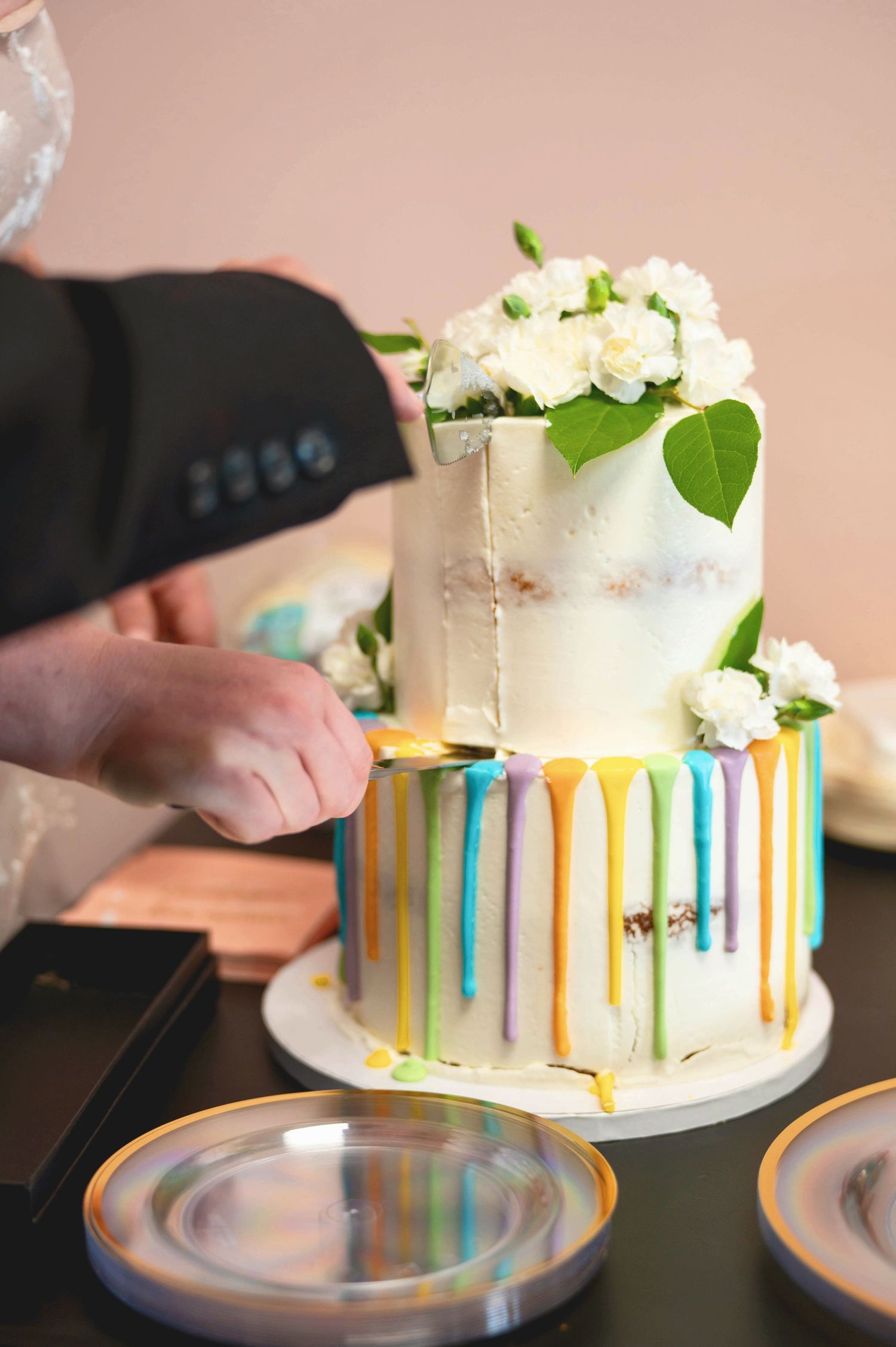 Two-tiered white wedding cake features colorful vertical stripes and floral decorations being cut at a reception table.
