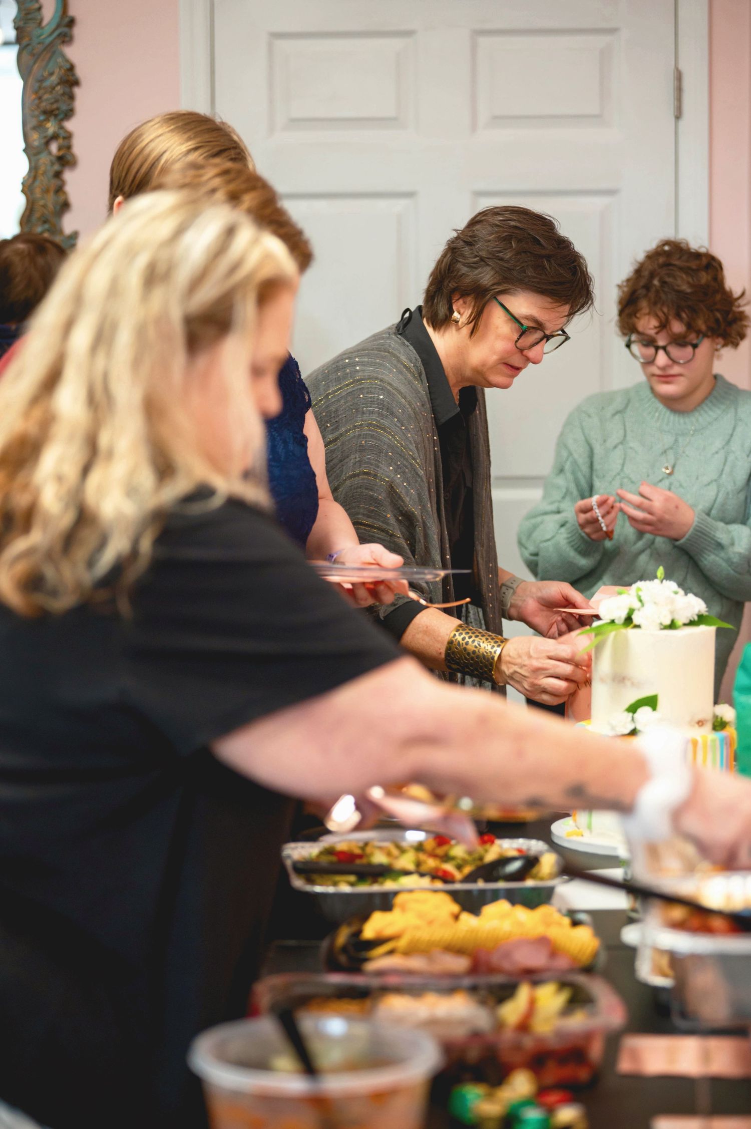 People gather around a dinner table serving themselves food from various dishes at what appears to be a casual gathering.