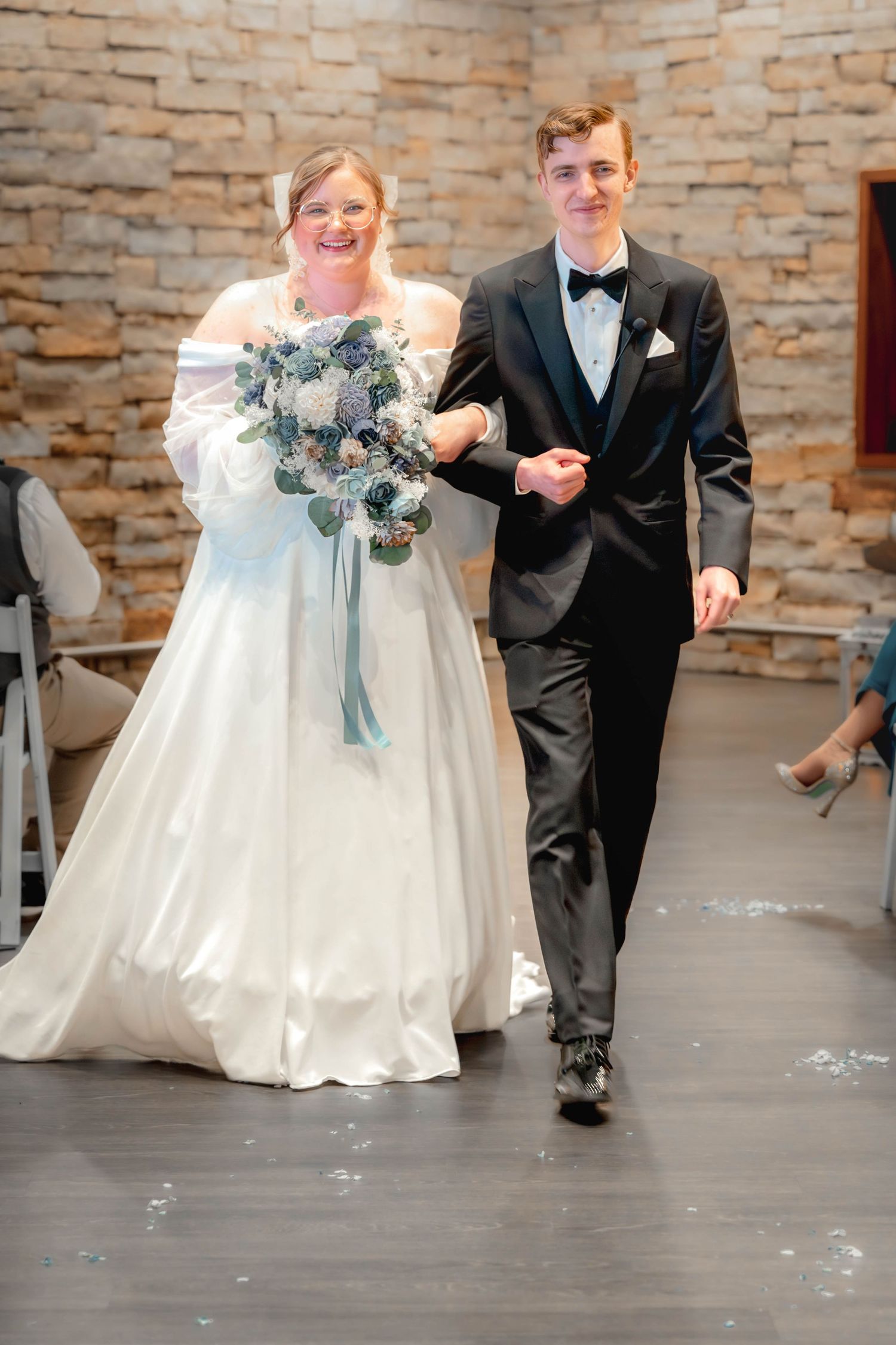A couple walks down the aisle at their wedding, with the bride wearing an off-shoulder gown and holding a blue-tinted bouquet.