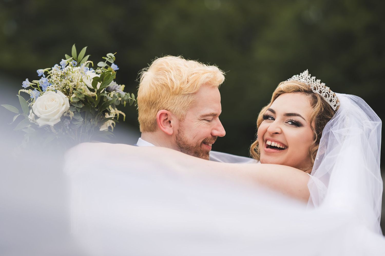 A joyful wedding couple shares a sweet moment together outdoors, with white flowers and greenery decorating the scene.